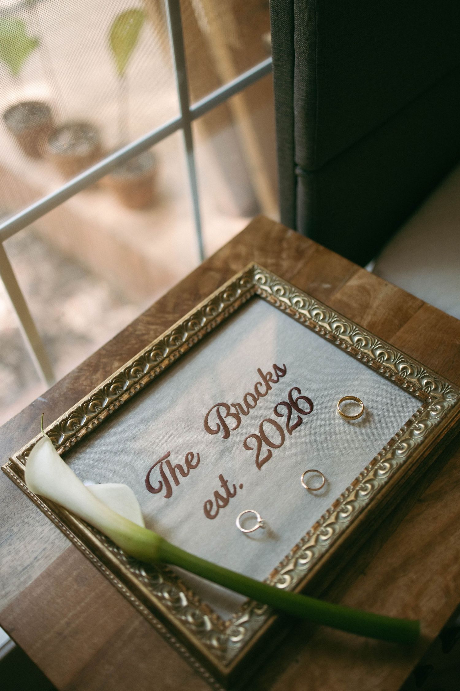 Gold-framed sign reading The Bracks est. 2026 with wedding rings and a white calla lily on a wooden surface.