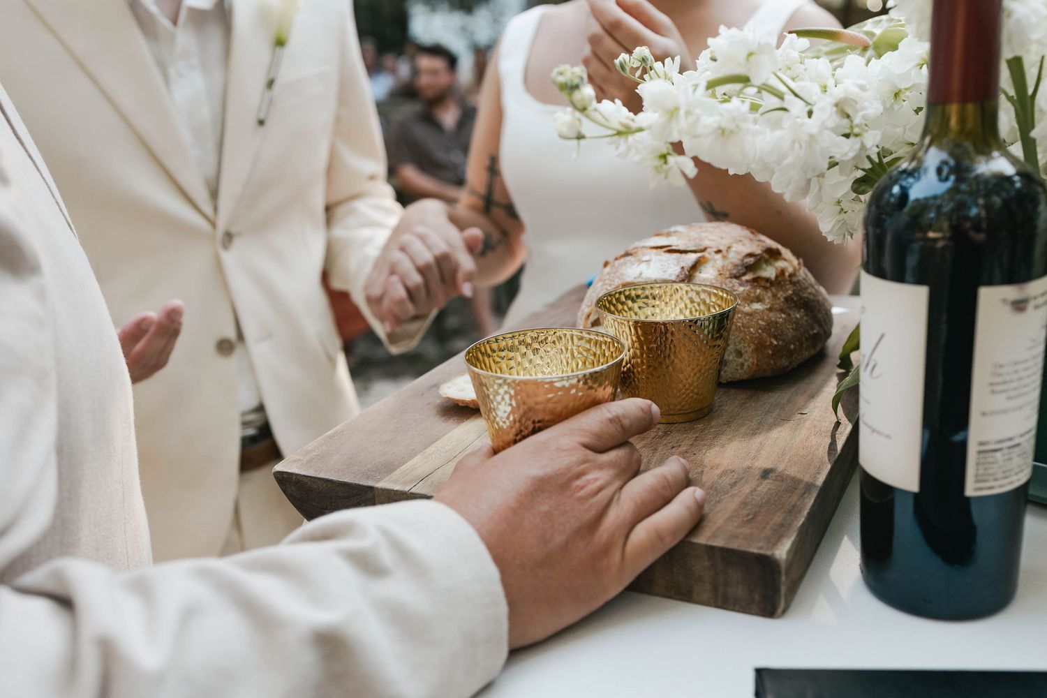Wedding ceremony table with bread, golden cups, and red wine bottle on wooden board.