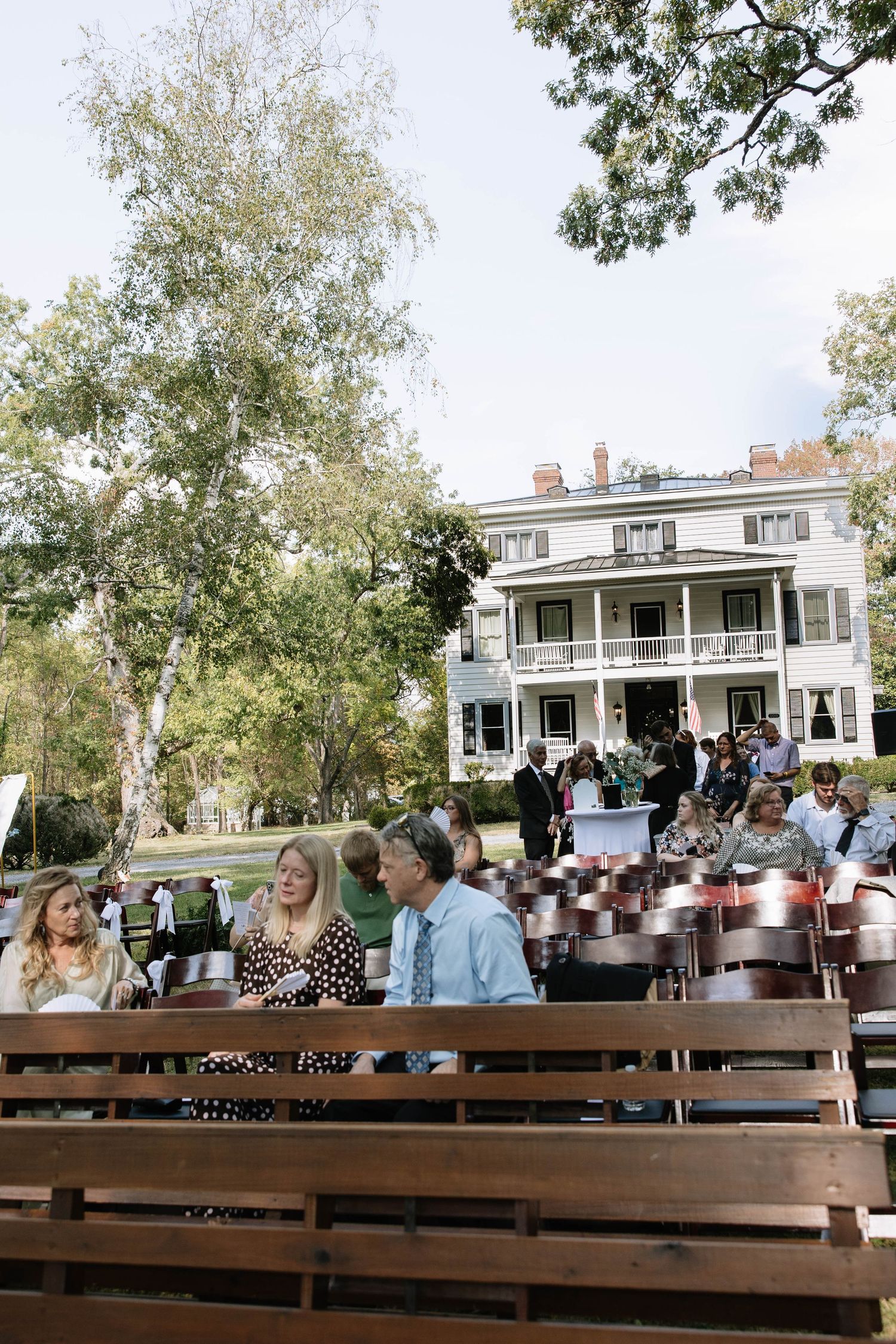 Wedding guests seated on wooden benches at an outdoor ceremony with a historic white house in the background.