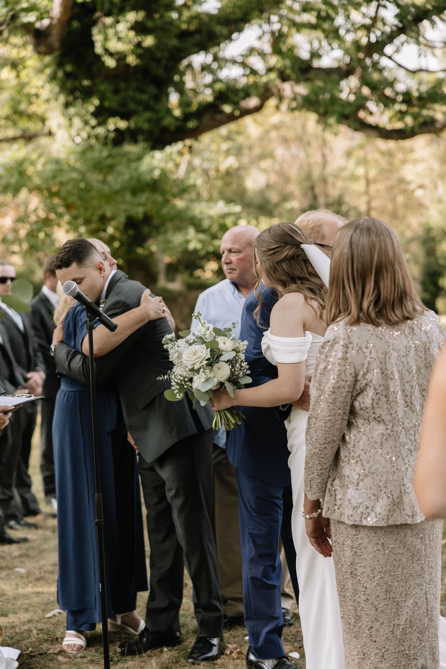 Wedding guests embrace and share emotional moments during an outdoor ceremony in a sunlit garden setting.