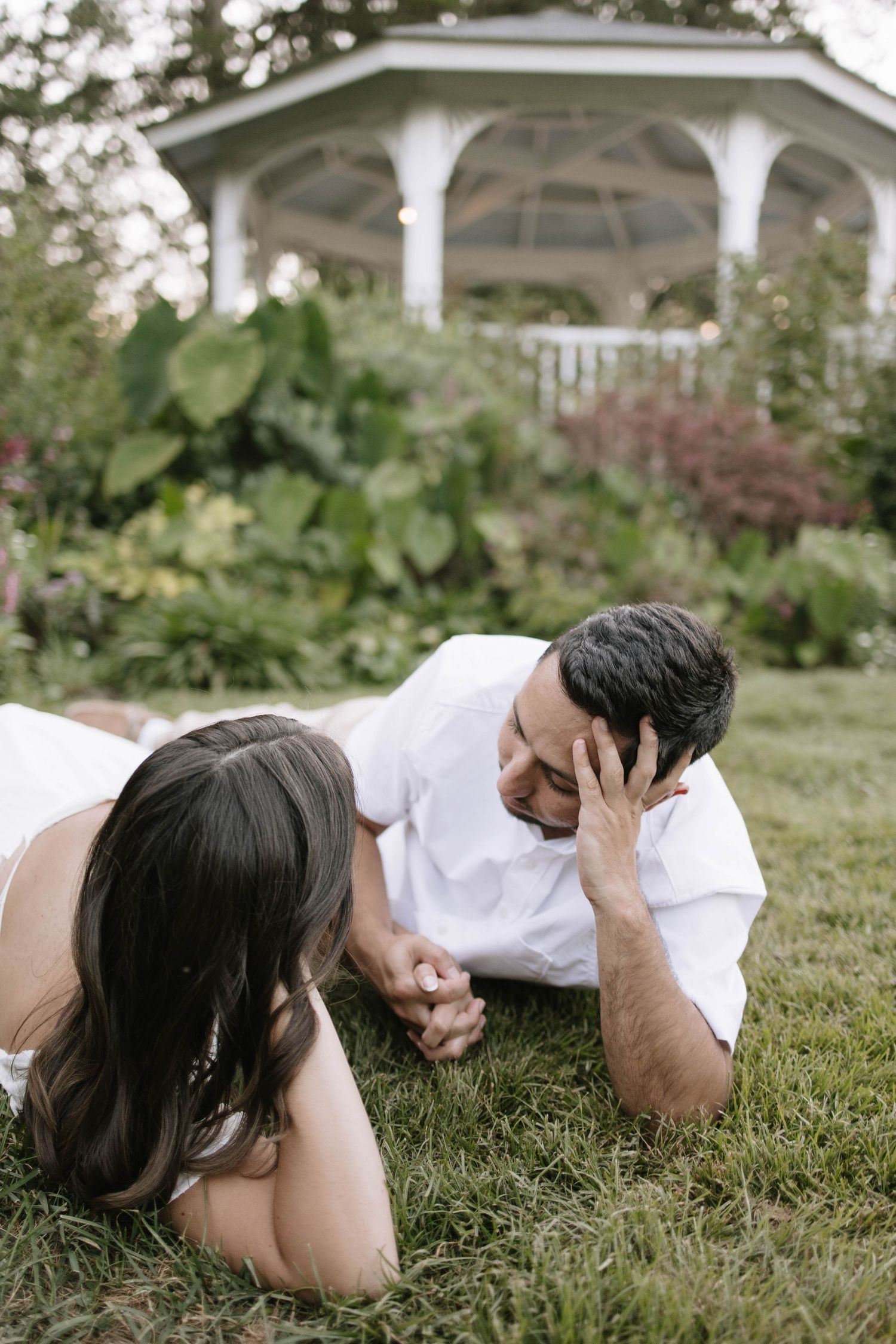 Couple lying in grass near white gazebo during romantic outdoor photoshoot.