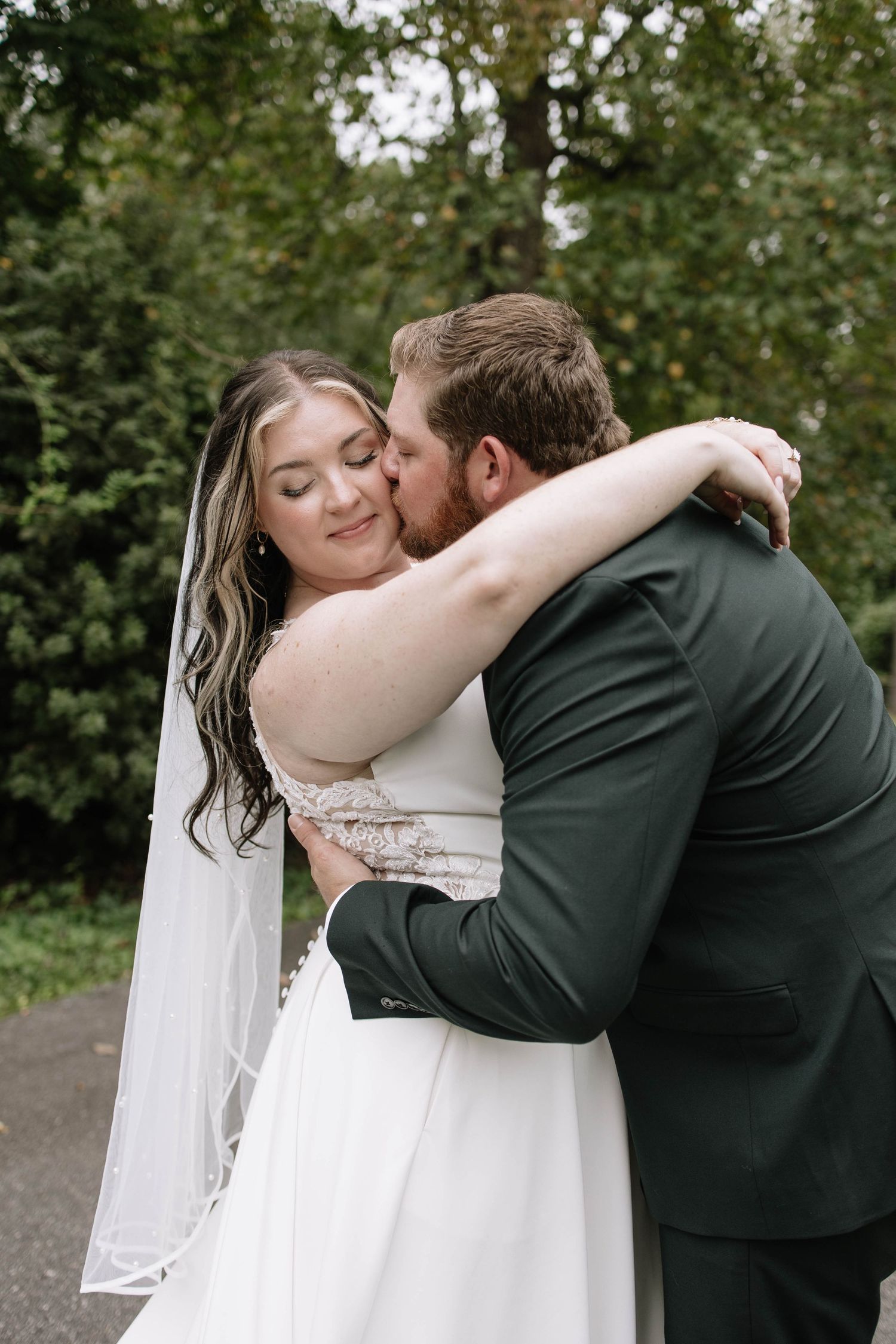 A romantic wedding couple embraces outdoors with the bride in white and groom in dark suit against a green backdrop.