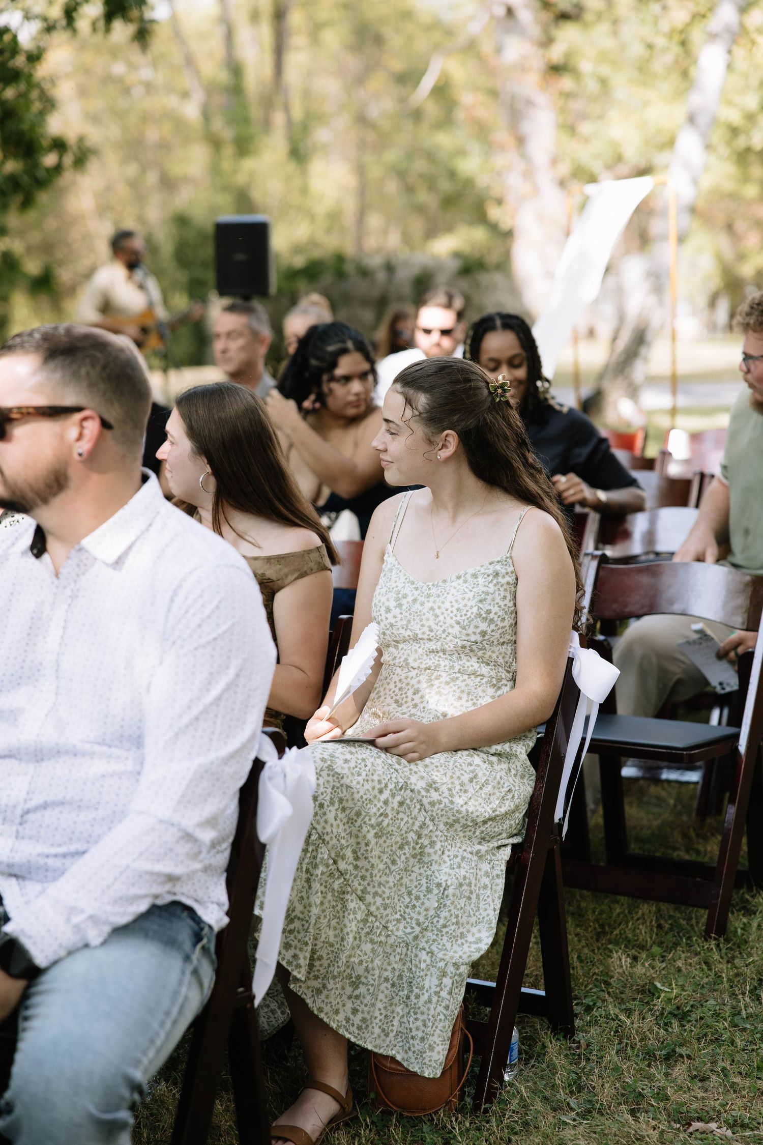 Wedding guests seated in wooden chairs at an outdoor ceremony under trees.