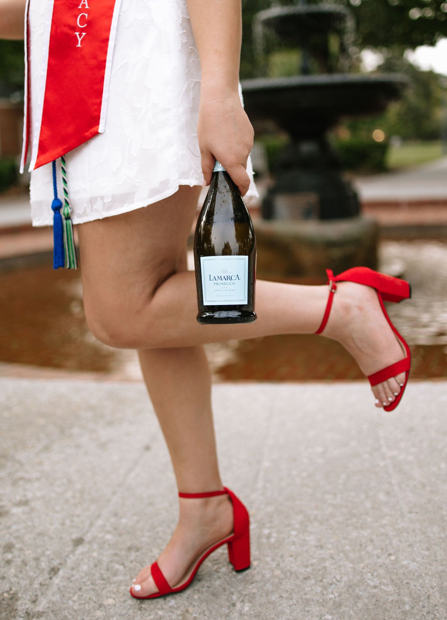 Graduate in white dress with red sash holds wine bottle, wearing red heels near fountain.