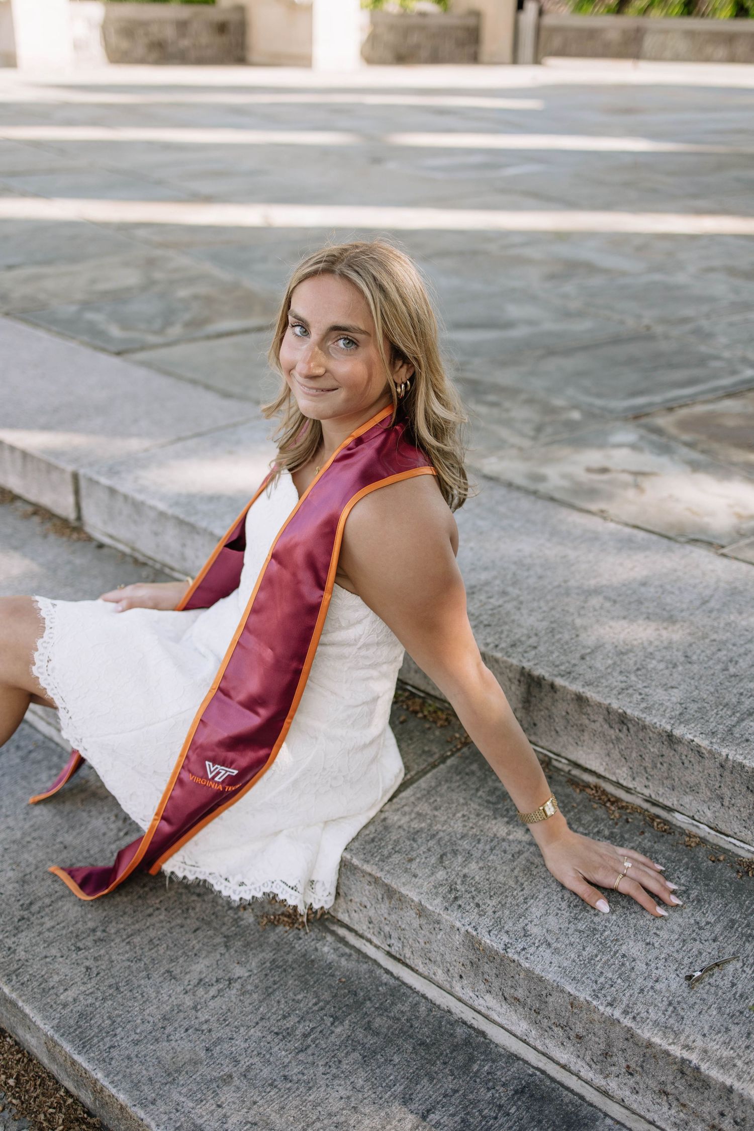 Young woman in white dress with maroon and gold Virginia Tech graduation sash sits on stone steps, smiling over shoulder.