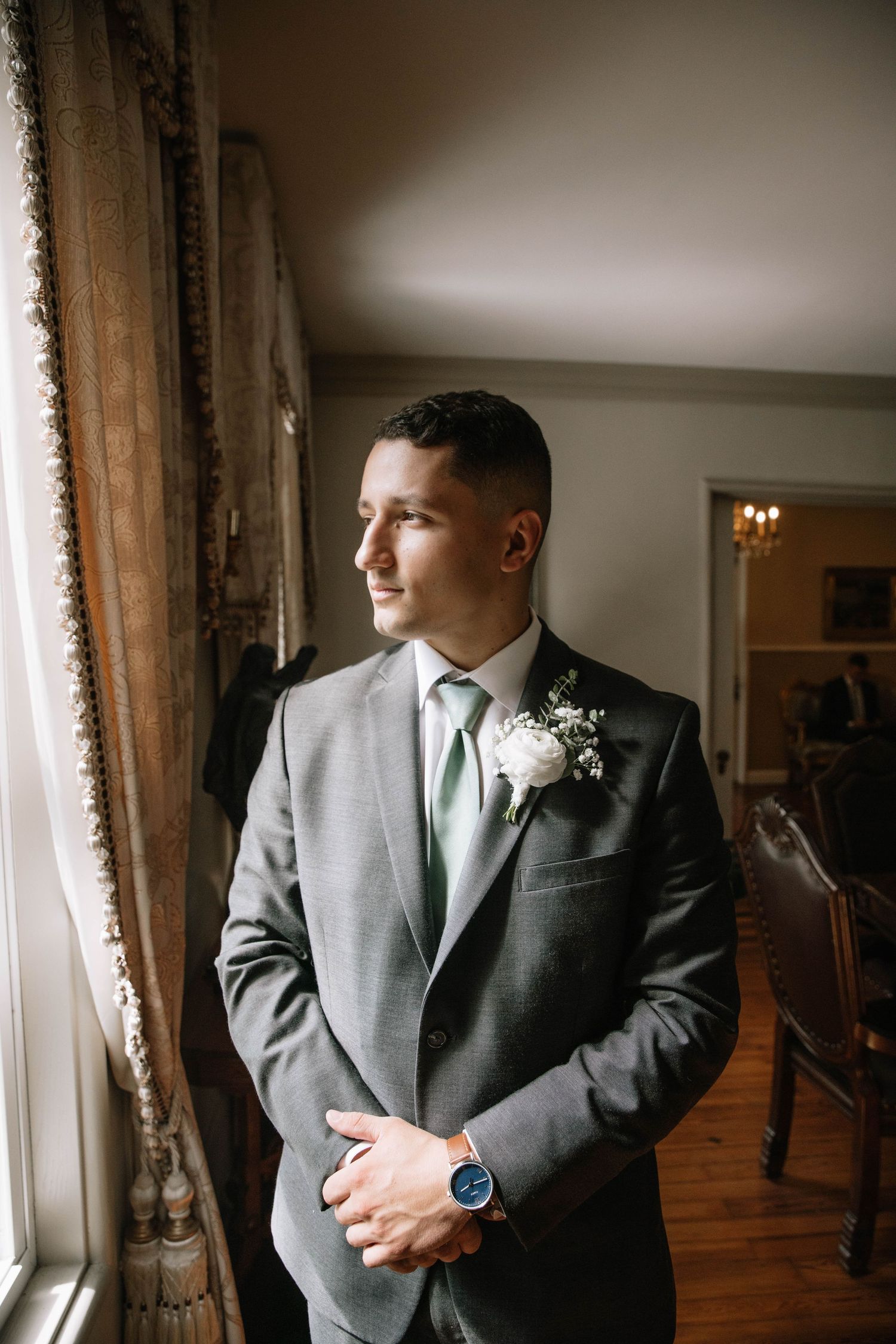 Groom in grey suit with mint tie stands thoughtfully by window in elegant venue with natural lighting.