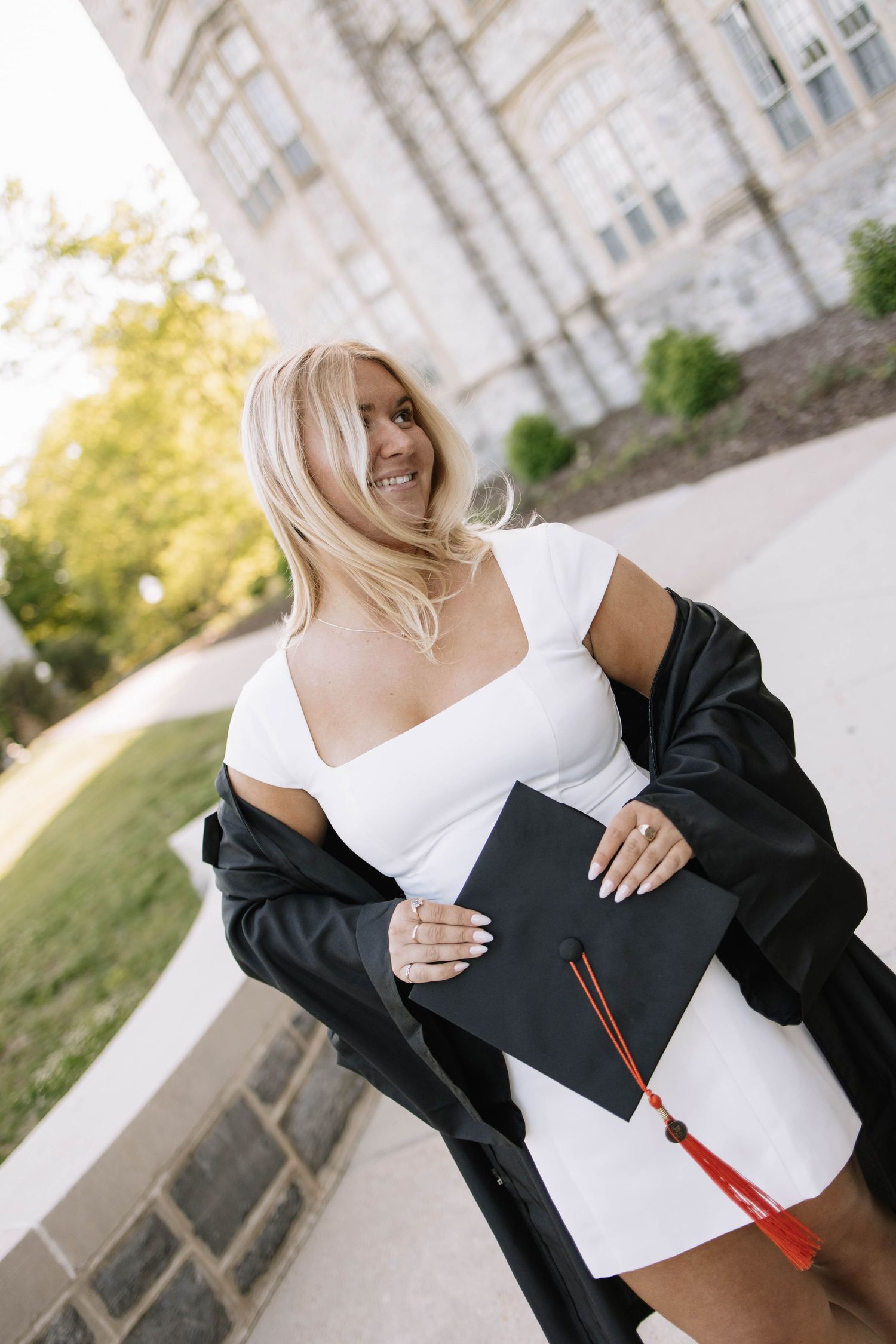 Smiling blonde graduate in white dress holding black cap with red tassel outside university building.