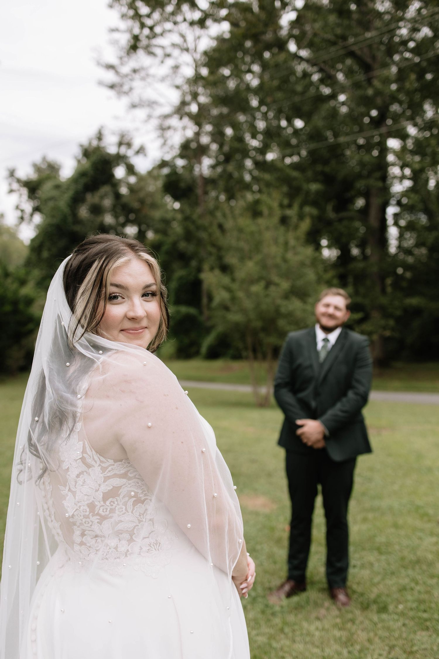 Bride in flowing white dress and veil poses playfully outdoors while groom stands in background.