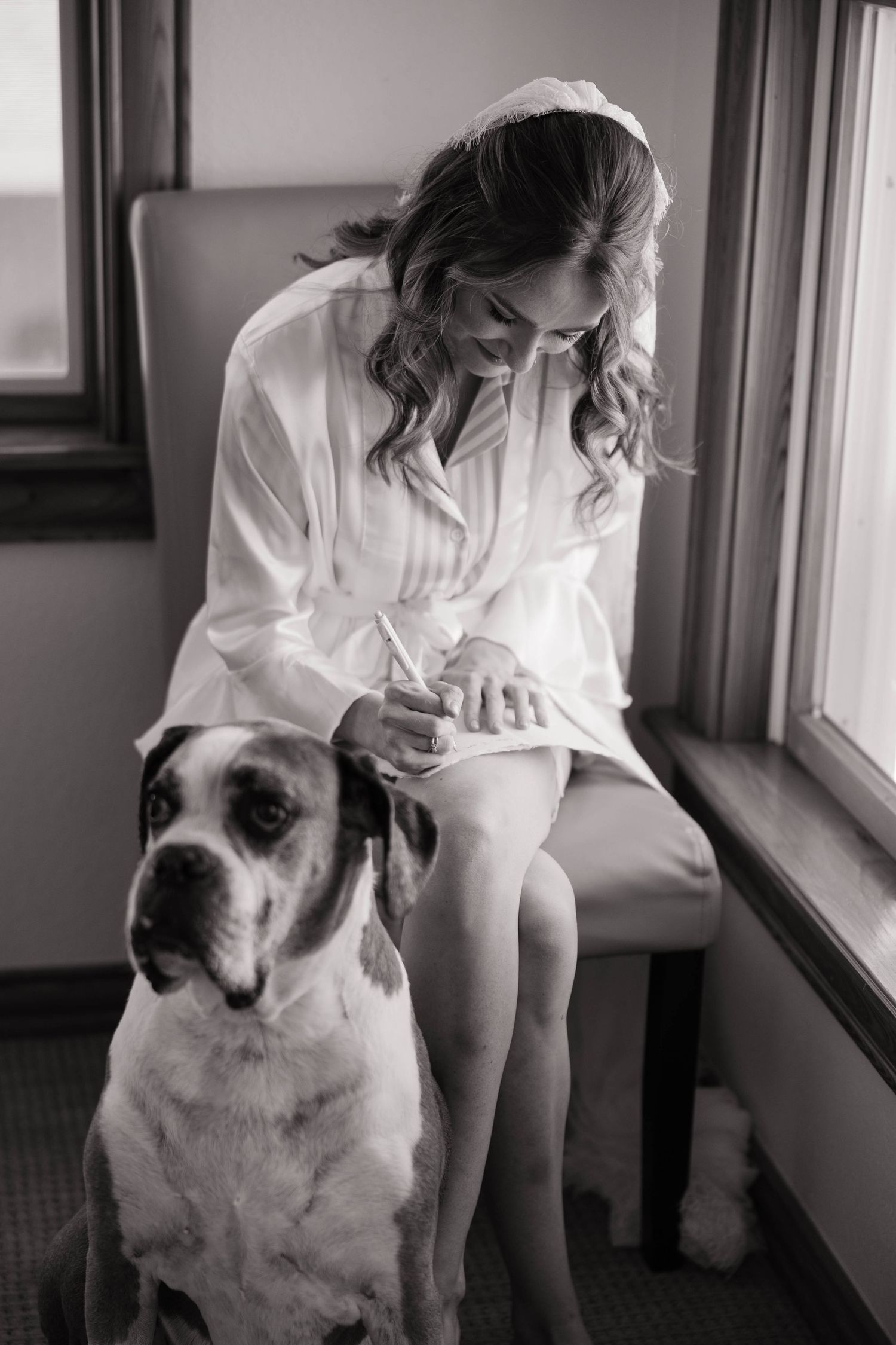 Black and white photo of a bride in white robe sitting by window with her Great Dane dog on her wedding day.