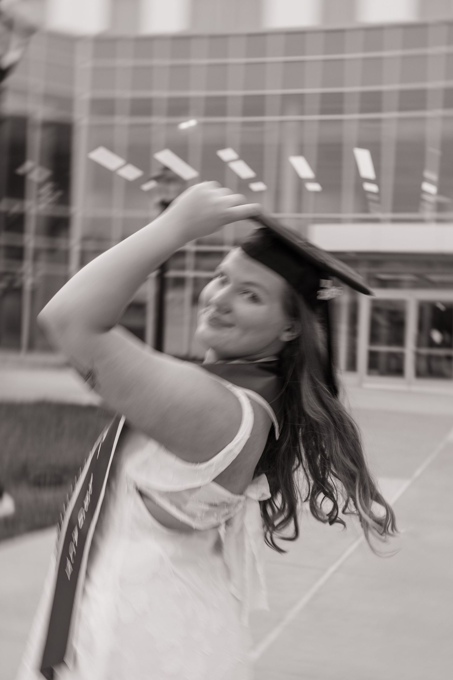 Black and white photo of a young woman in graduation cap and gown, joyfully posing outdoors near a modern building.