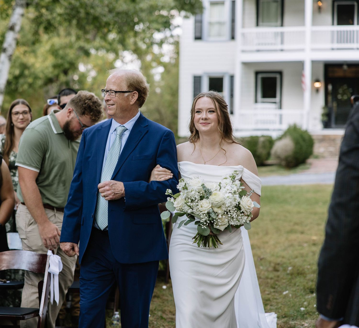 Guests gather outdoors at an elegant wedding ceremony with white buildings and green lawns in the background.