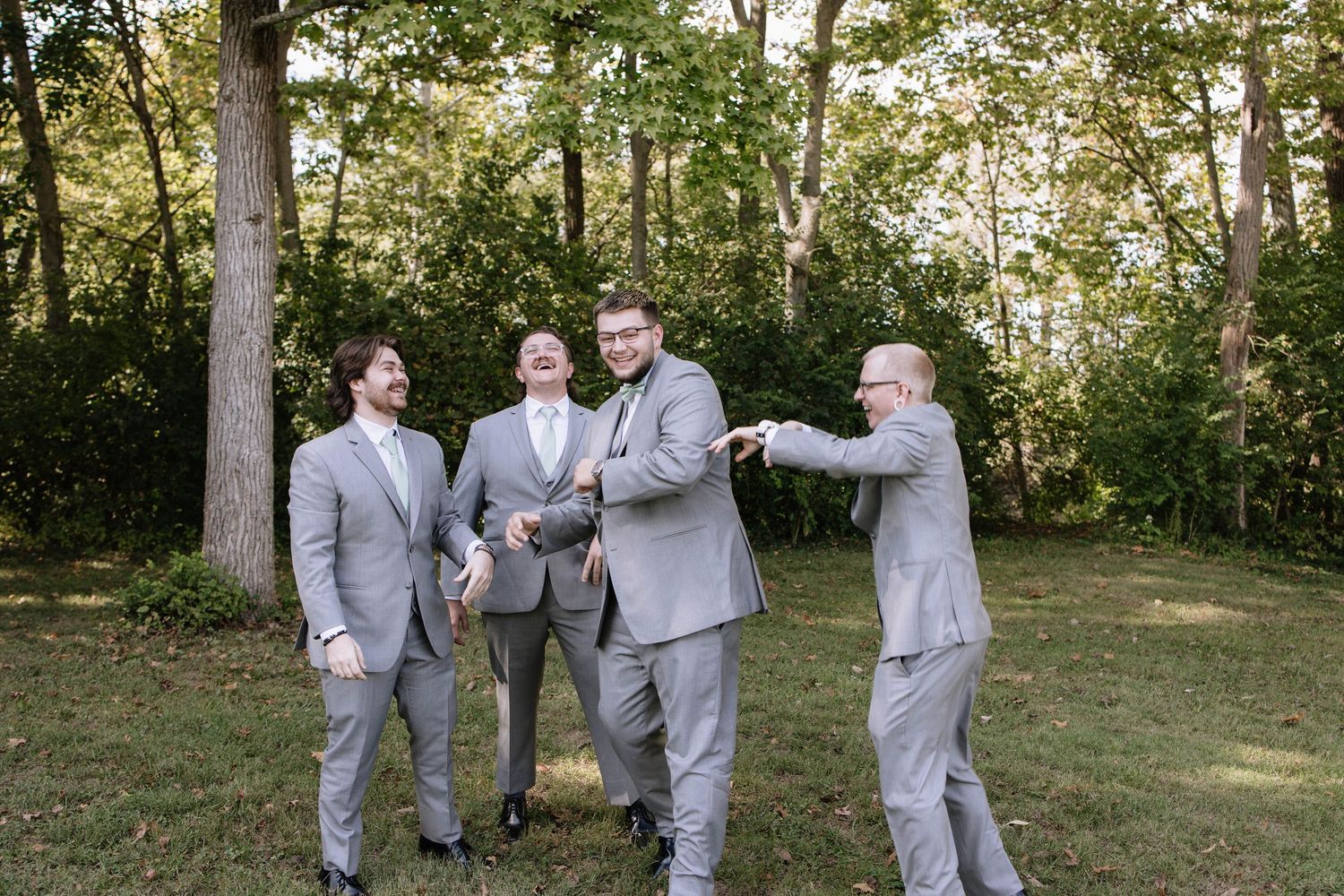 Groomsmen in light grey suits share a laugh during wedding party photos outdoors.