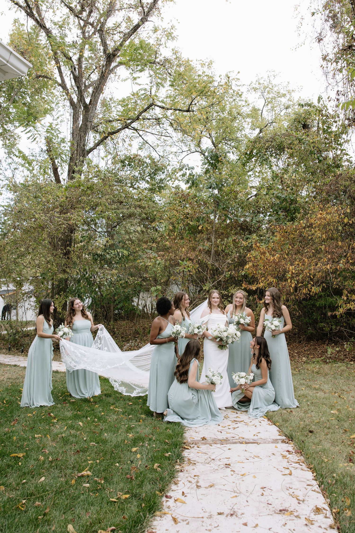 Bridesmaids in light blue dresses help arrange the bride's wedding gown outdoors among autumn trees.