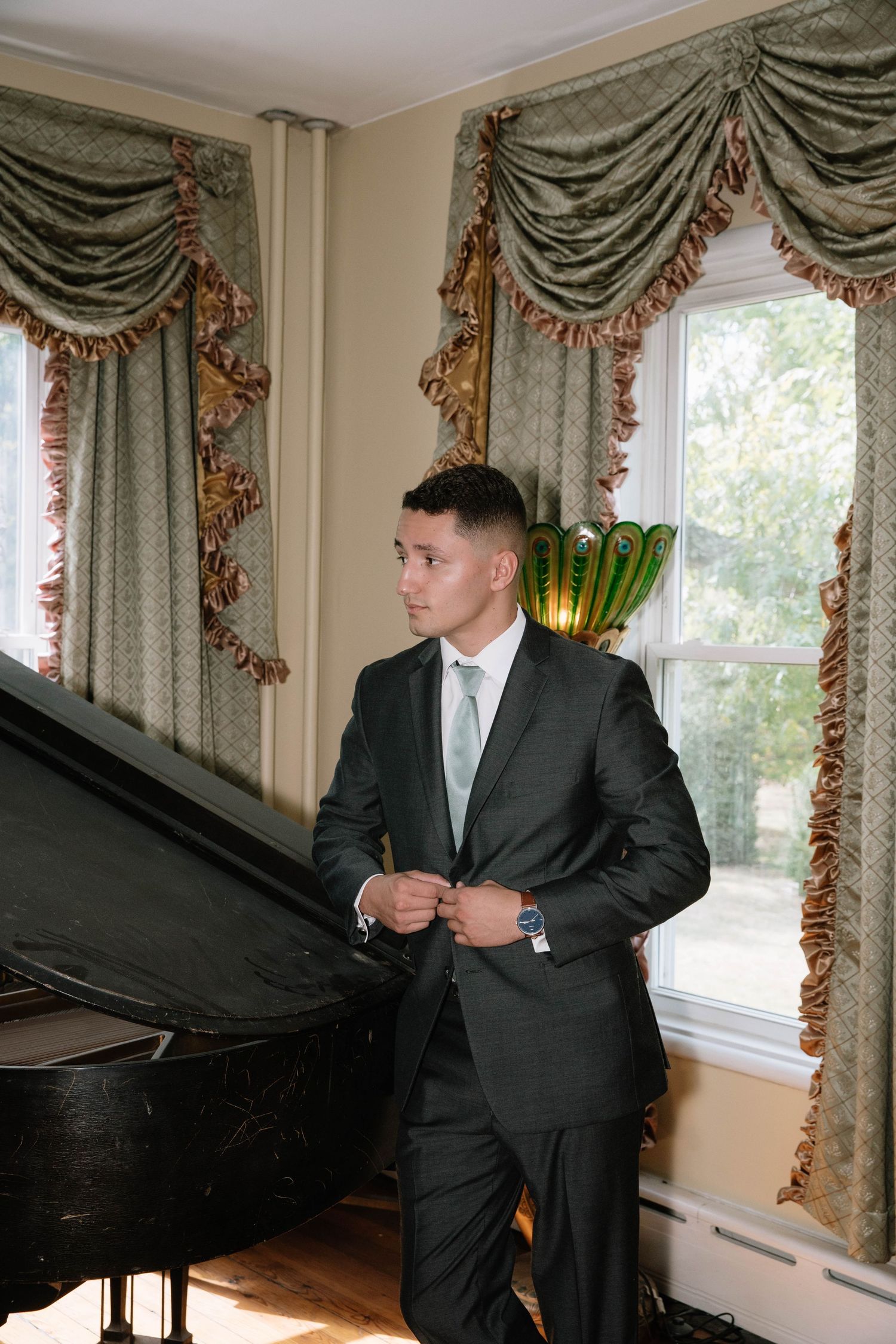 A person in a dark suit stands beside an elegant black grand piano in a room with ornate sage green curtains.