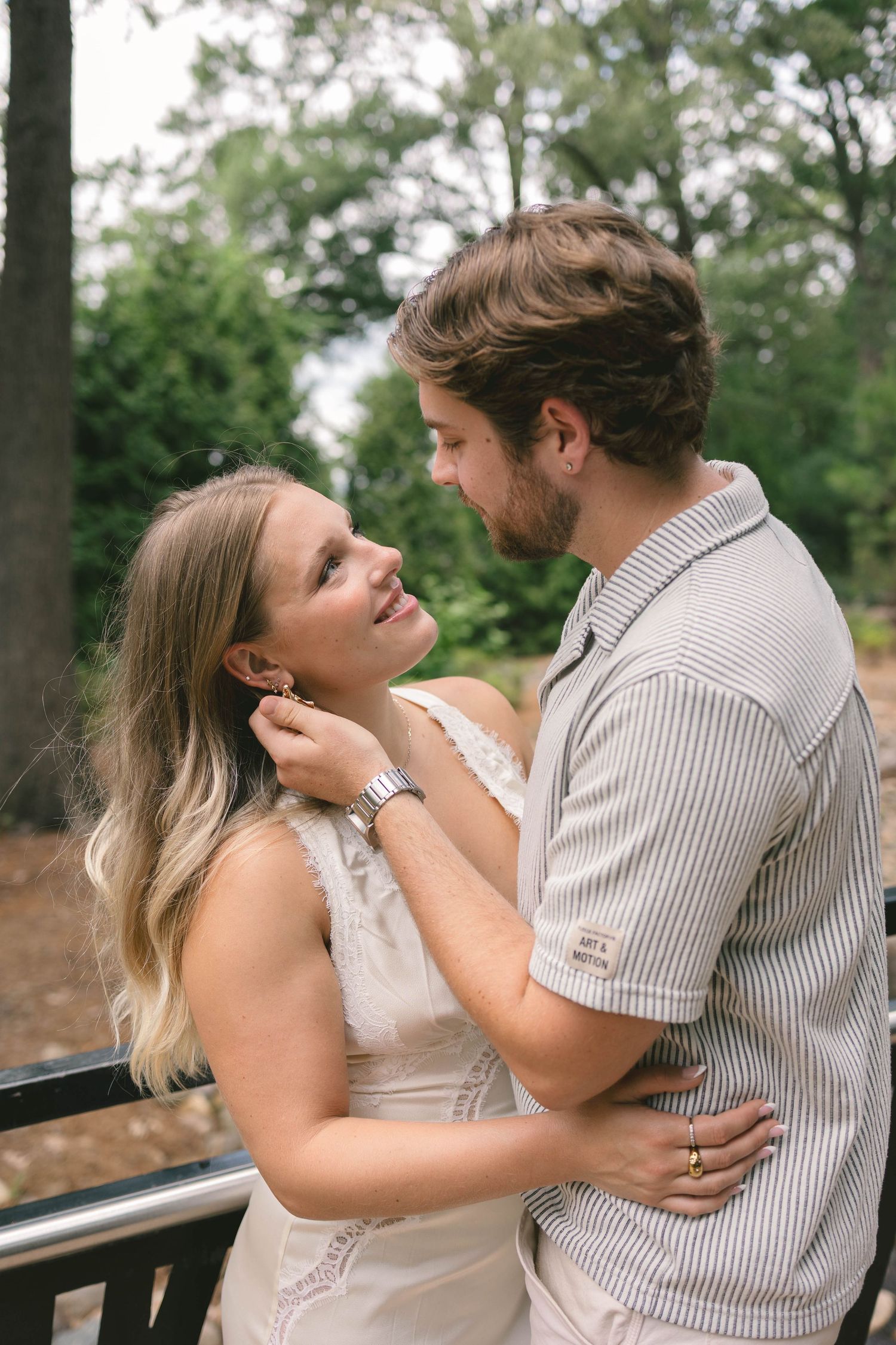 A couple shares a tender moment outdoors, as a man gently touches a woman's hair while she smiles up at him.