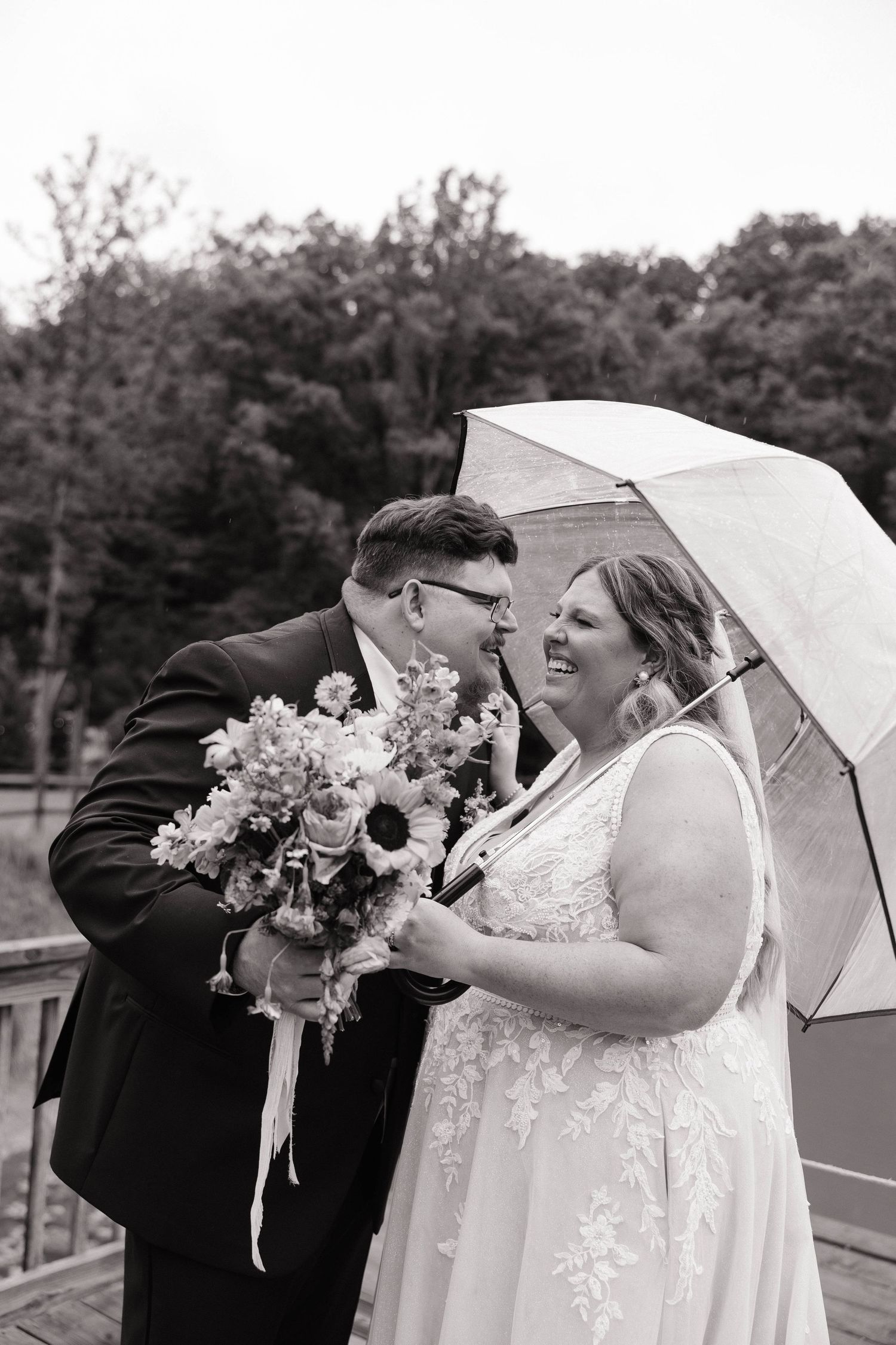 Black and white photo of a smiling bride and groom sharing a moment under a clear umbrella holding a floral bouquet.