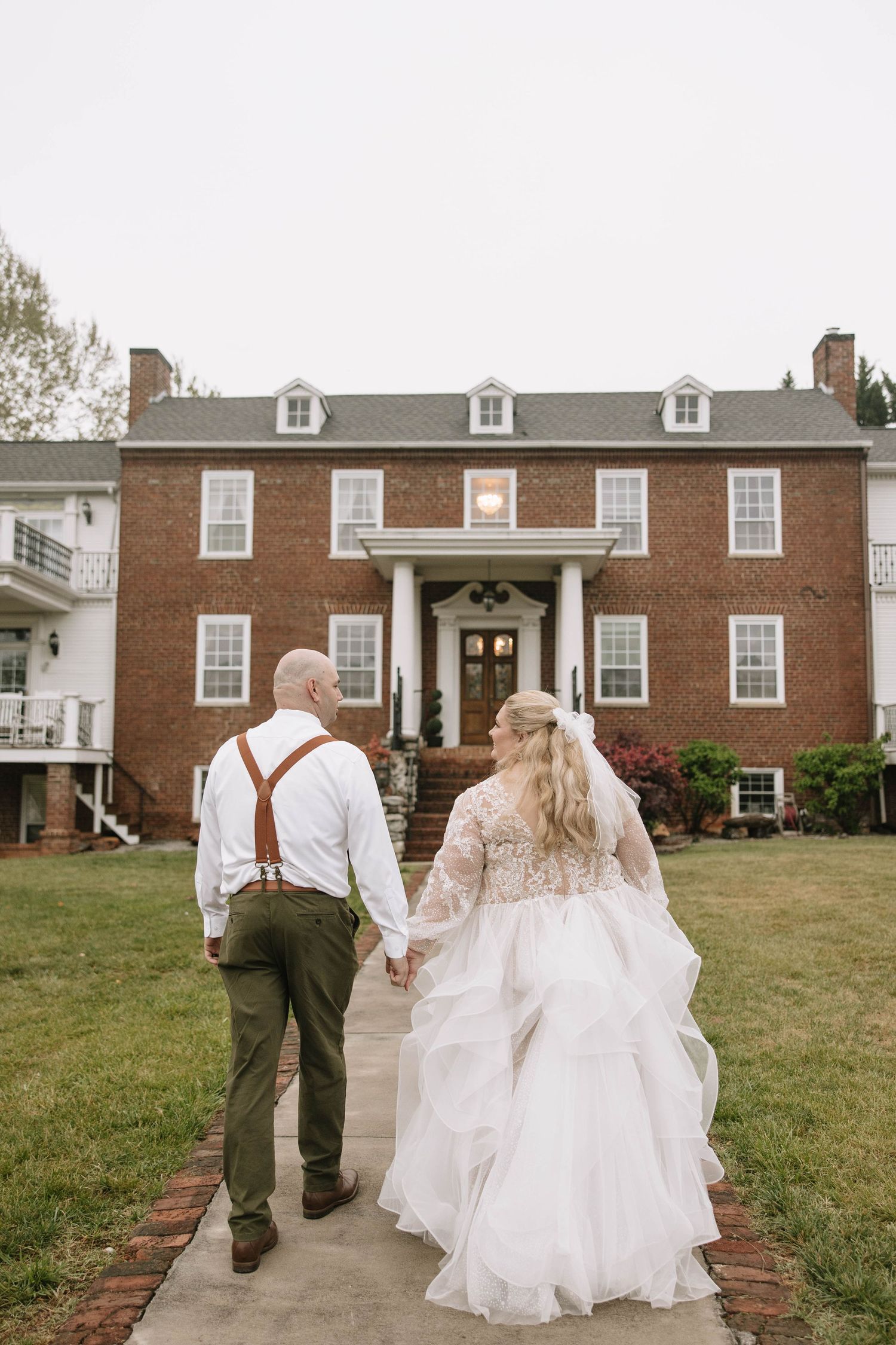Bride and groom walking hand in hand toward a grand brick colonial mansion on their wedding day.