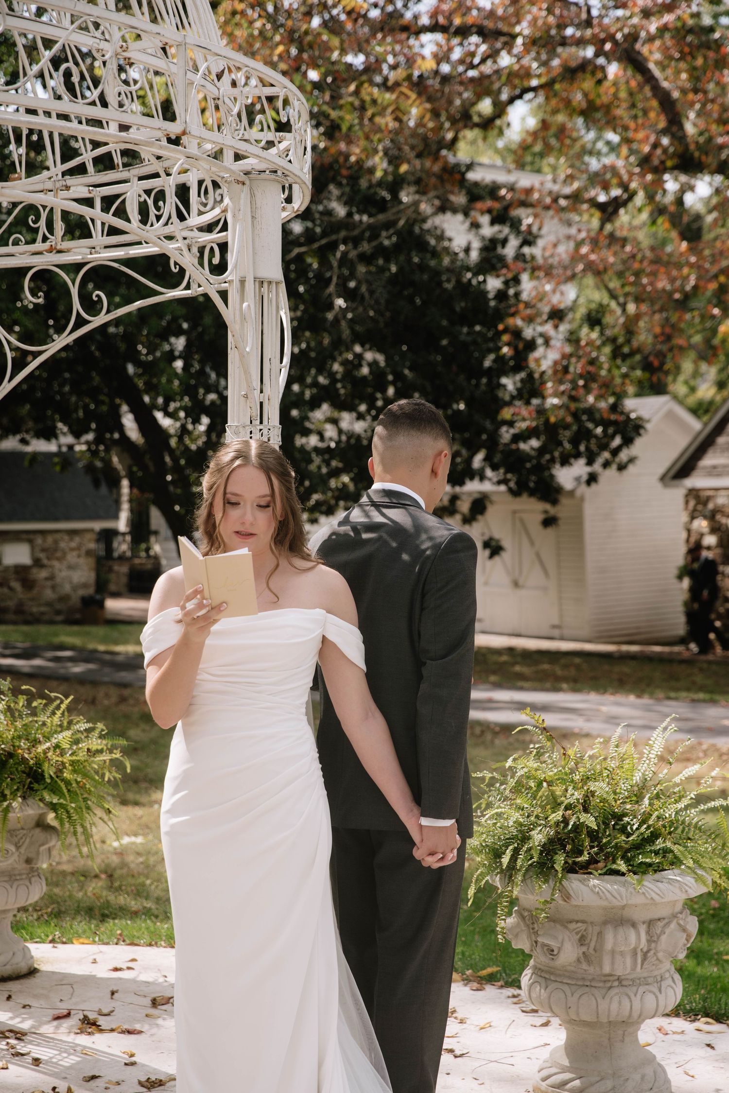 Elegant wedding couple poses together under a white ornate gazebo surrounded by autumn foliage in garden setting.