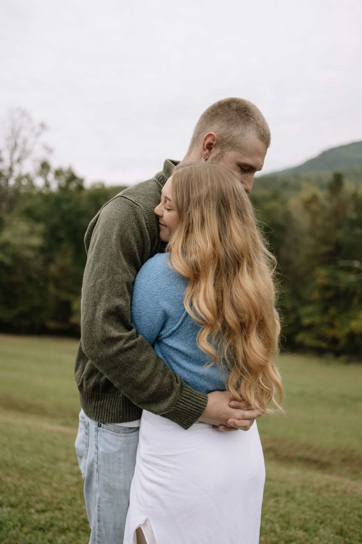 A romantic embrace between a couple in casual attire against a rural mountain backdrop.