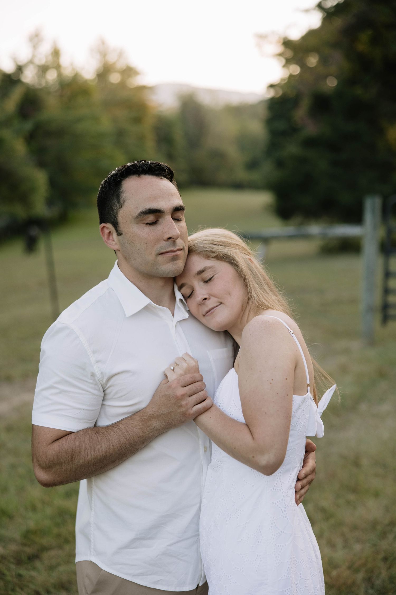 A romantic couple in white outfits embraces tenderly during a sunset photoshoot in a grassy field.