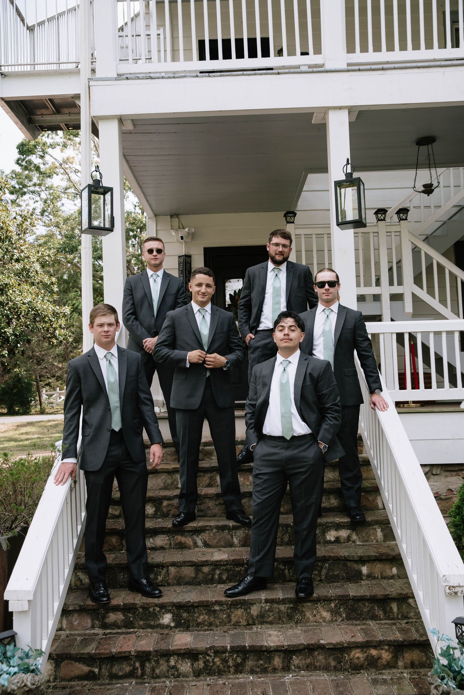 Group of groomsmen in formal attire relaxing on stone steps of a white building.