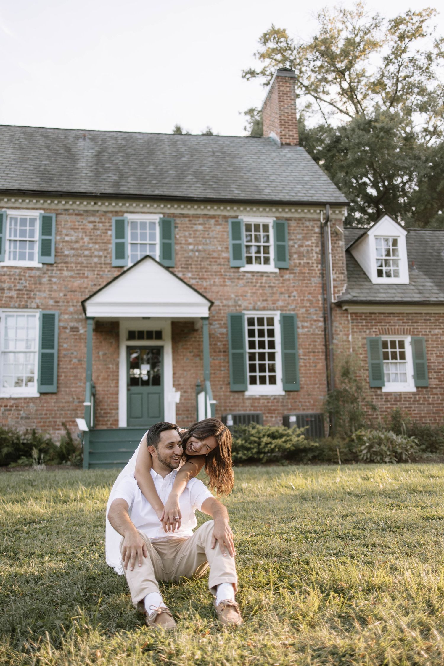A couple poses playfully in front of a historic brick house with white trim and green shutters.