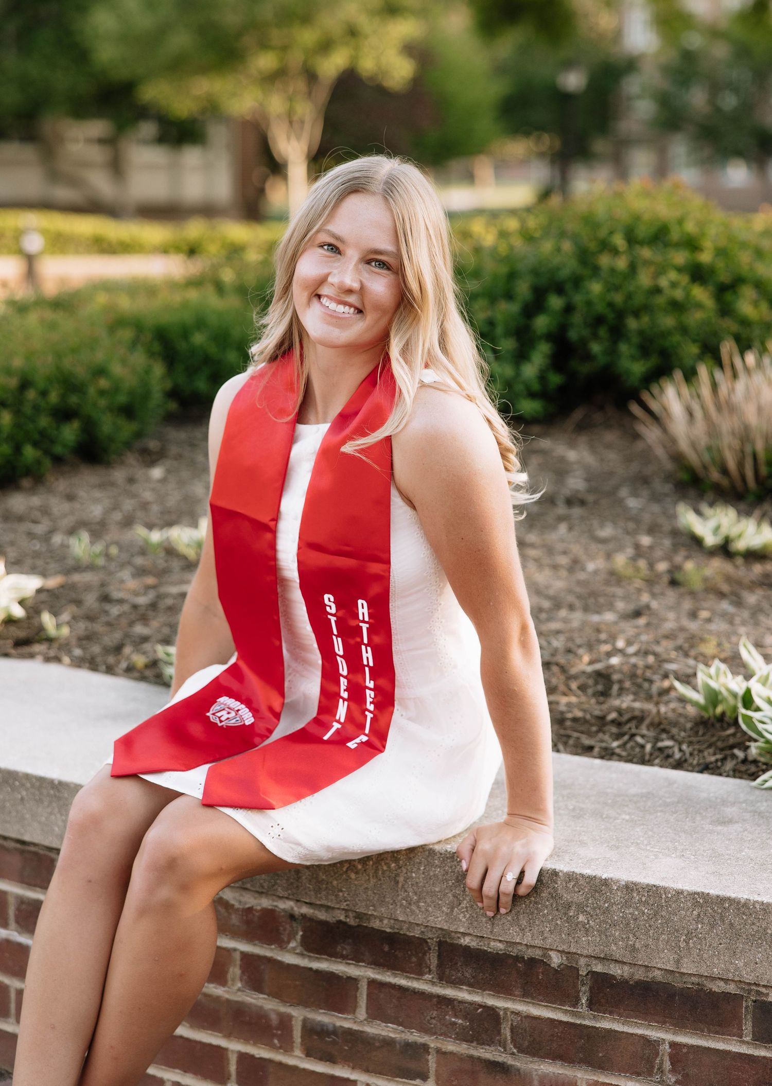 Smiling blonde woman in white dress wearing red graduation stole seated outdoors on brick wall at university campus.