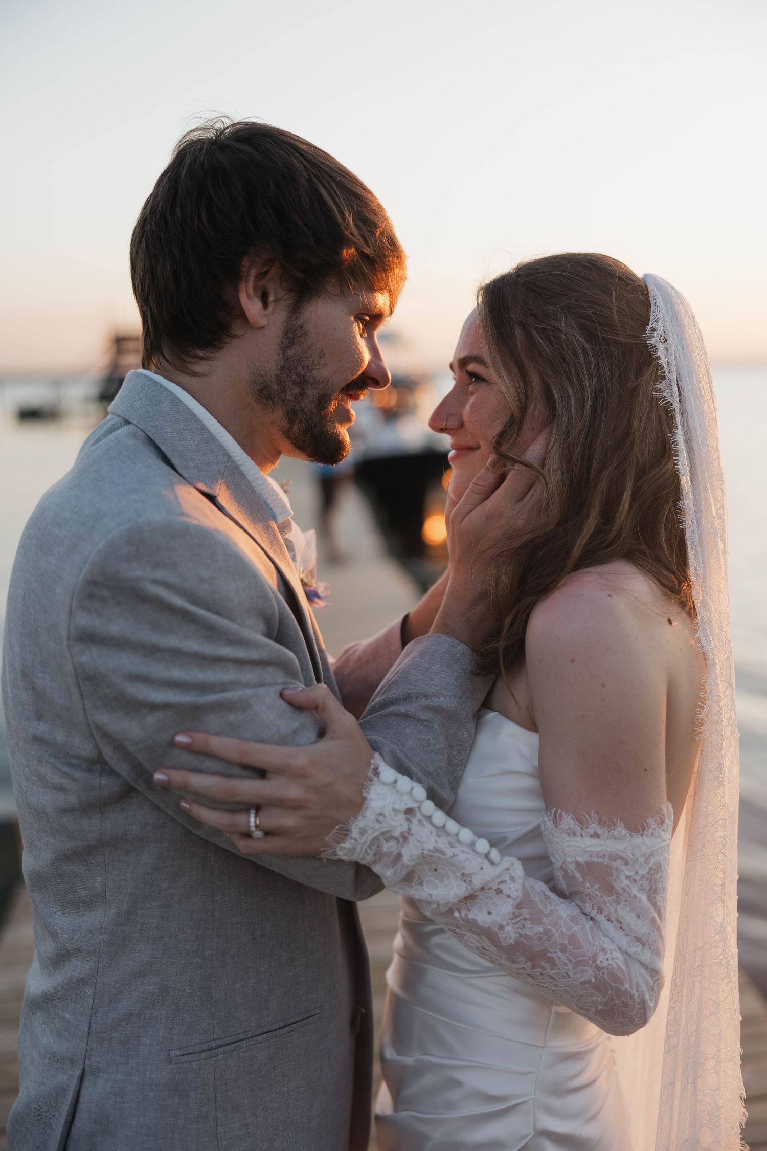 A bride and groom share a romantic moment at sunset by the water, with the groom tenderly cupping the bride's face.