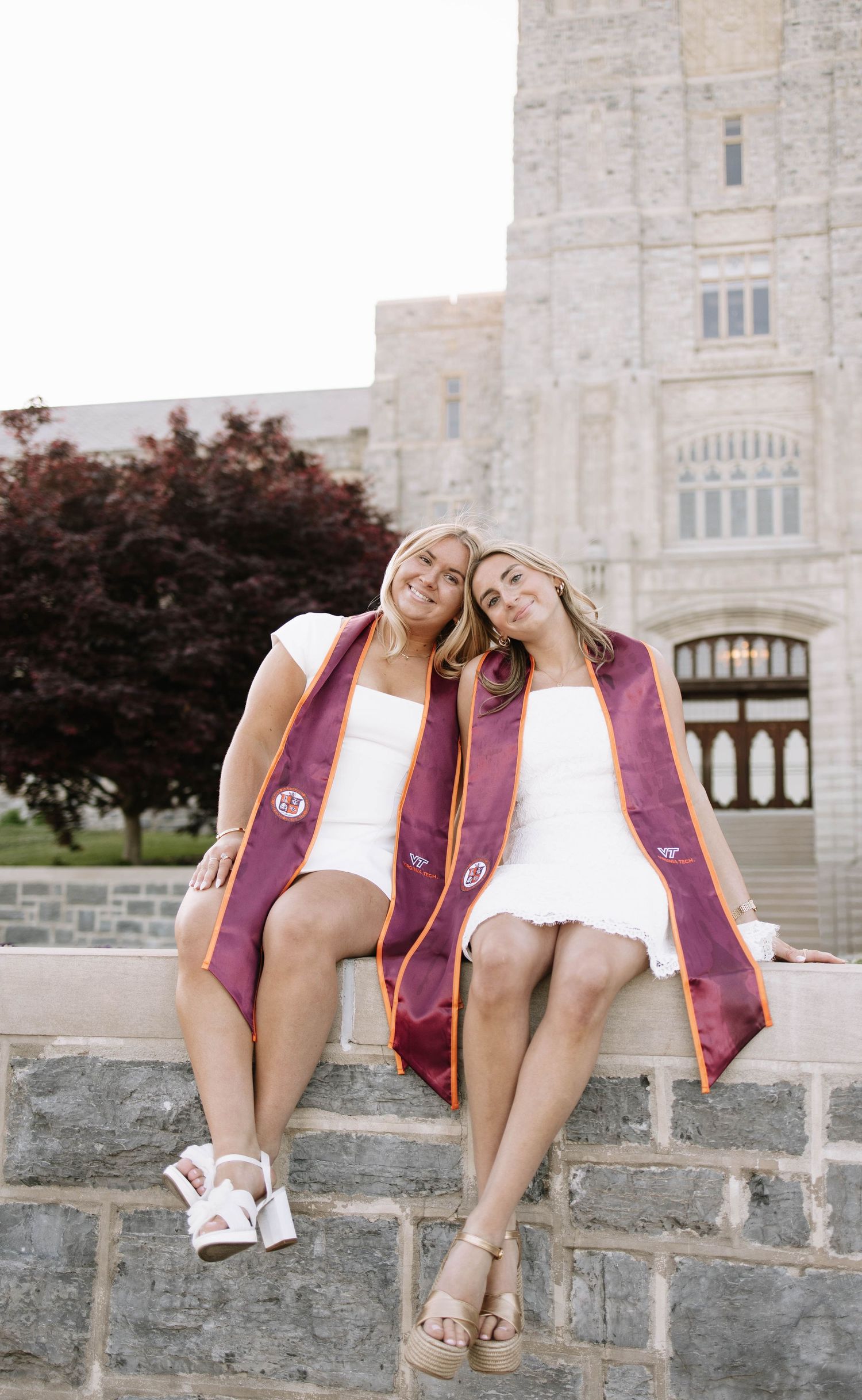 Two smiling graduates in white dresses wear maroon stoles while sitting on a stone wall outside a university building.