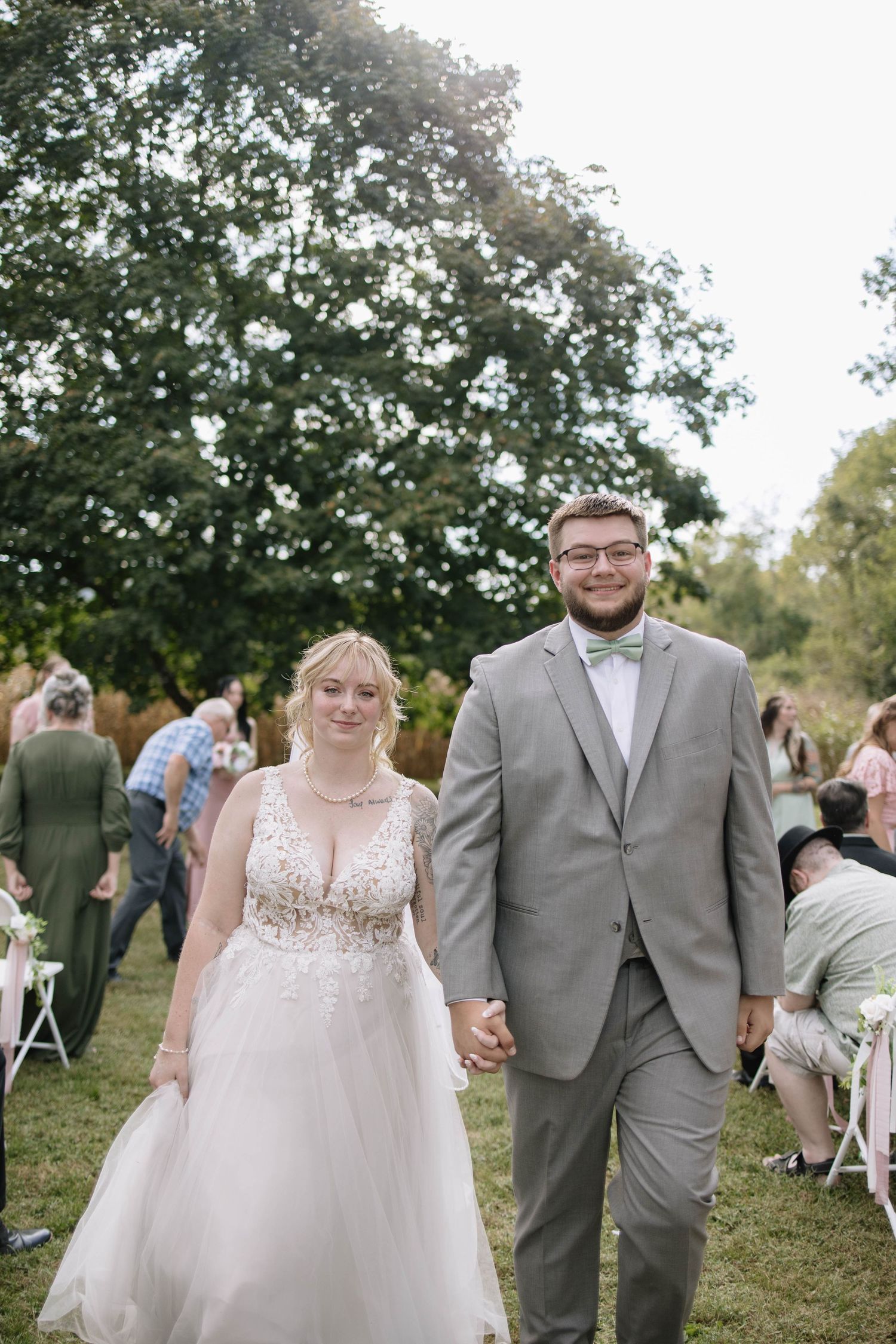 Wedding couple walking hand in hand outdoors with guests visible in background.