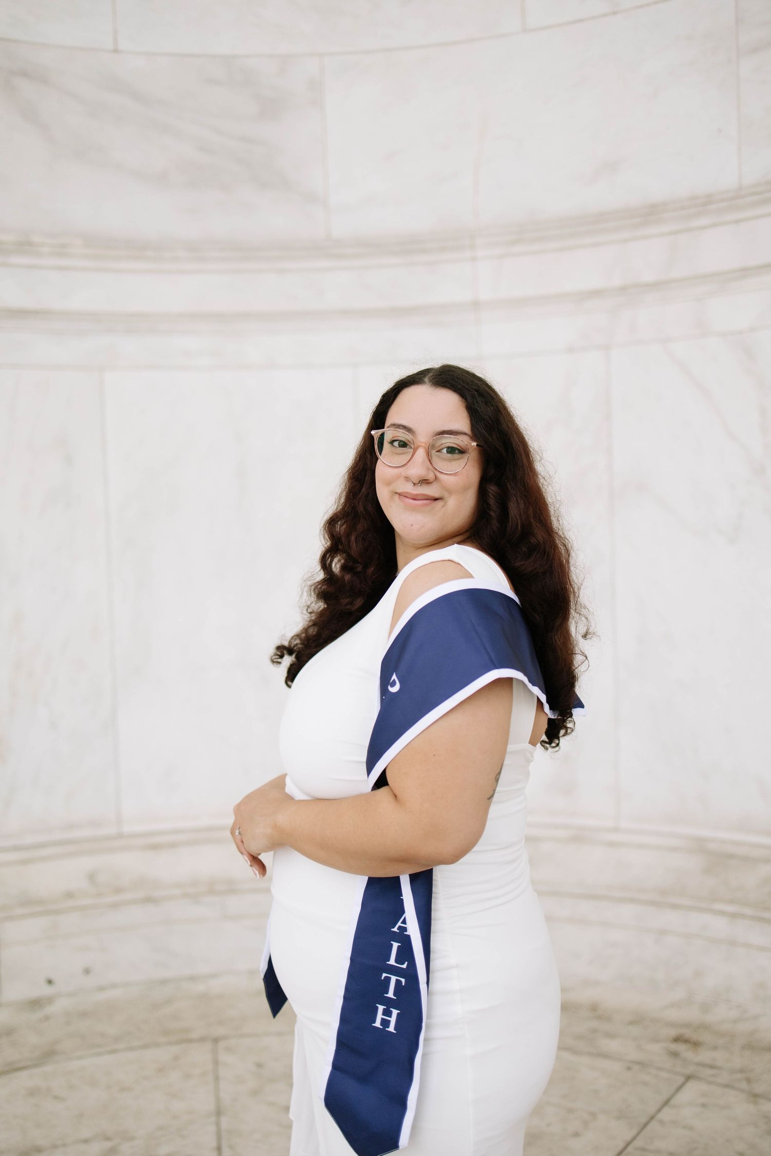 Young woman in white dress with navy blue graduation sash smiles over shoulder at marble monument.