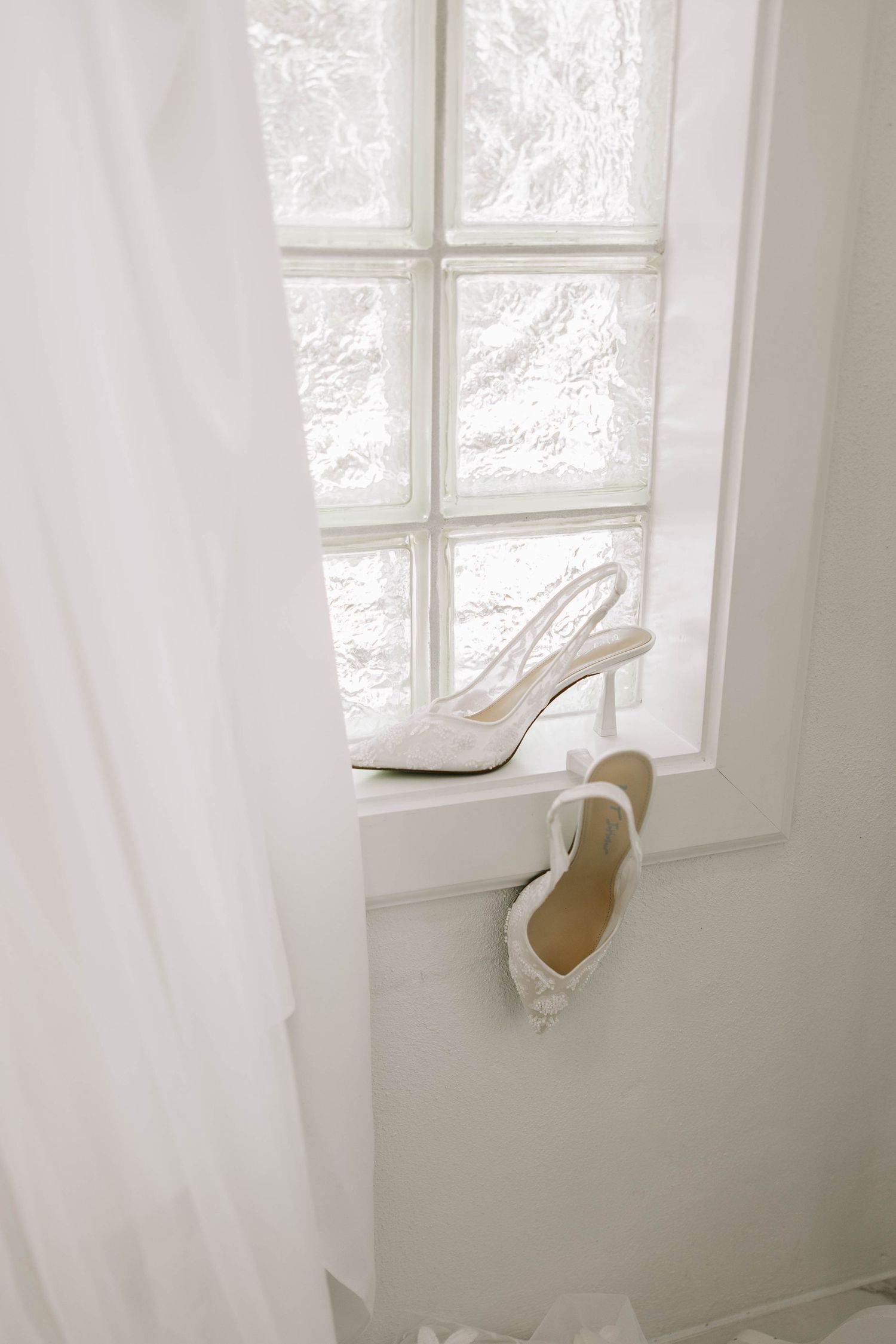 Elegant white slingback heels displayed on a windowsill with sheer curtains and natural light streaming through.
