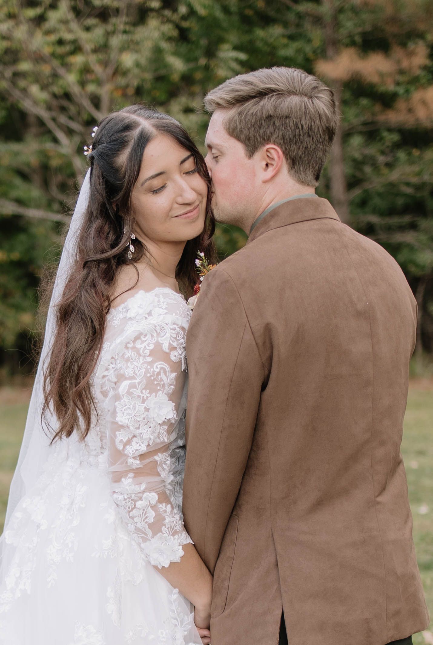 Bride in lace gown embraces groom in brown suit during outdoor portraits.