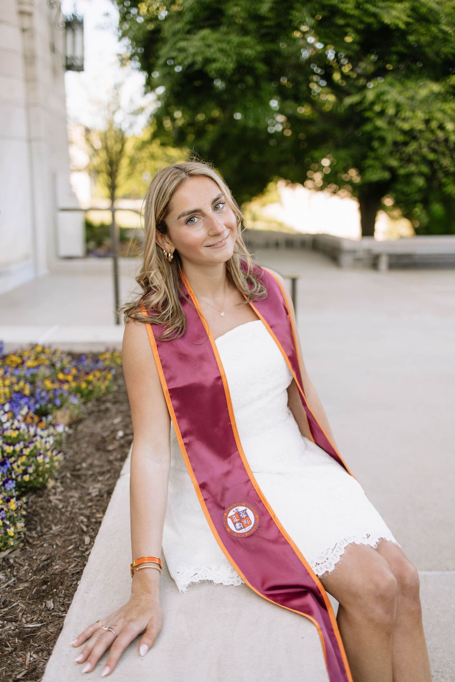 Young woman in white dress wearing maroon graduation sash sits outdoors on stone ledge surrounded by greenery.