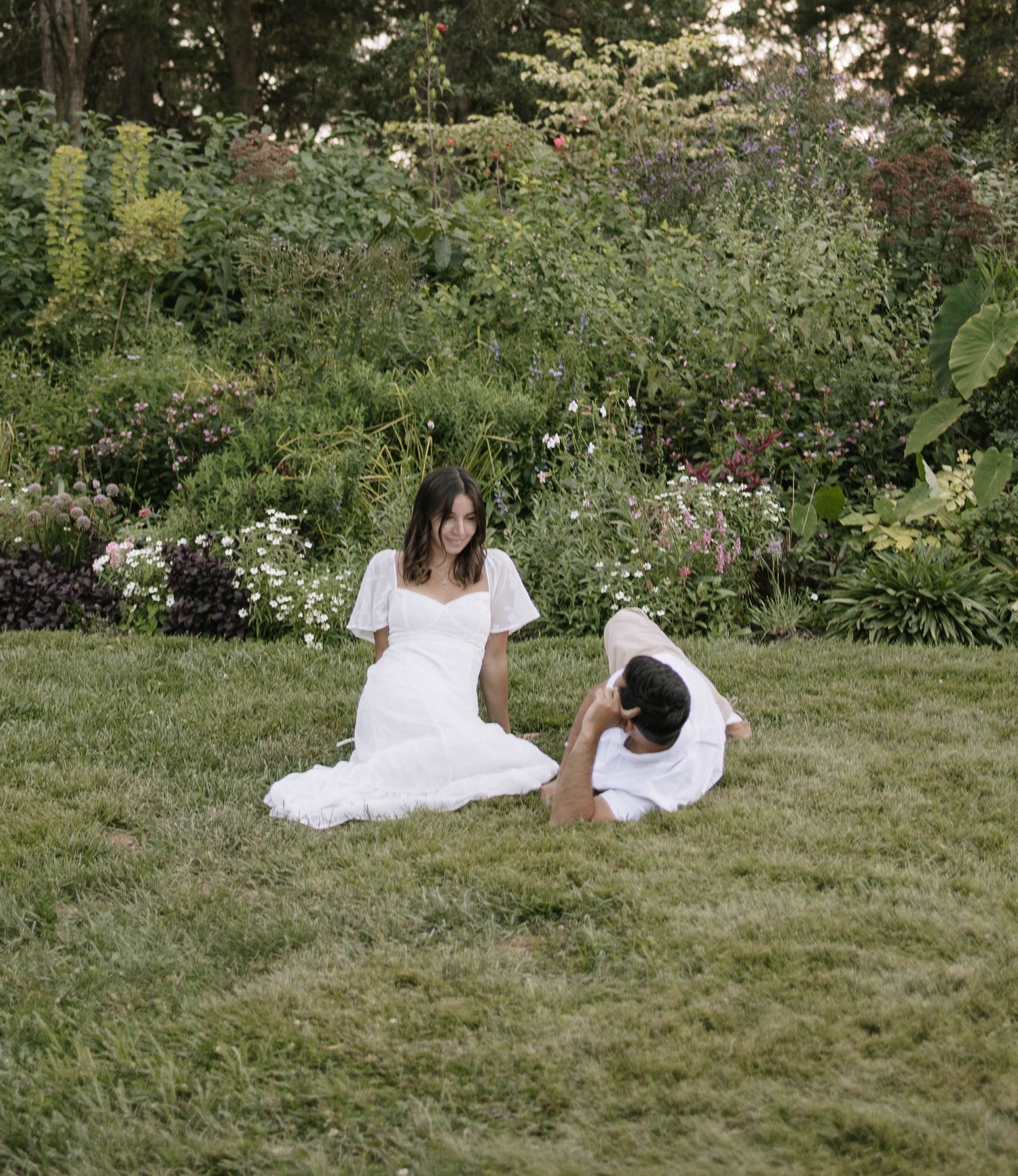 Relaxed couple lounges on lawn surrounded by garden flowers during summer photo session.