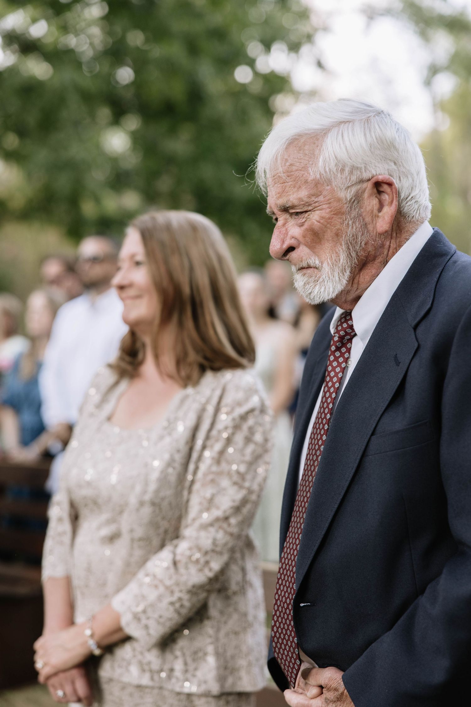 Elderly man in dark suit and tie stands next to woman in sparkly dress at outdoor gathering.