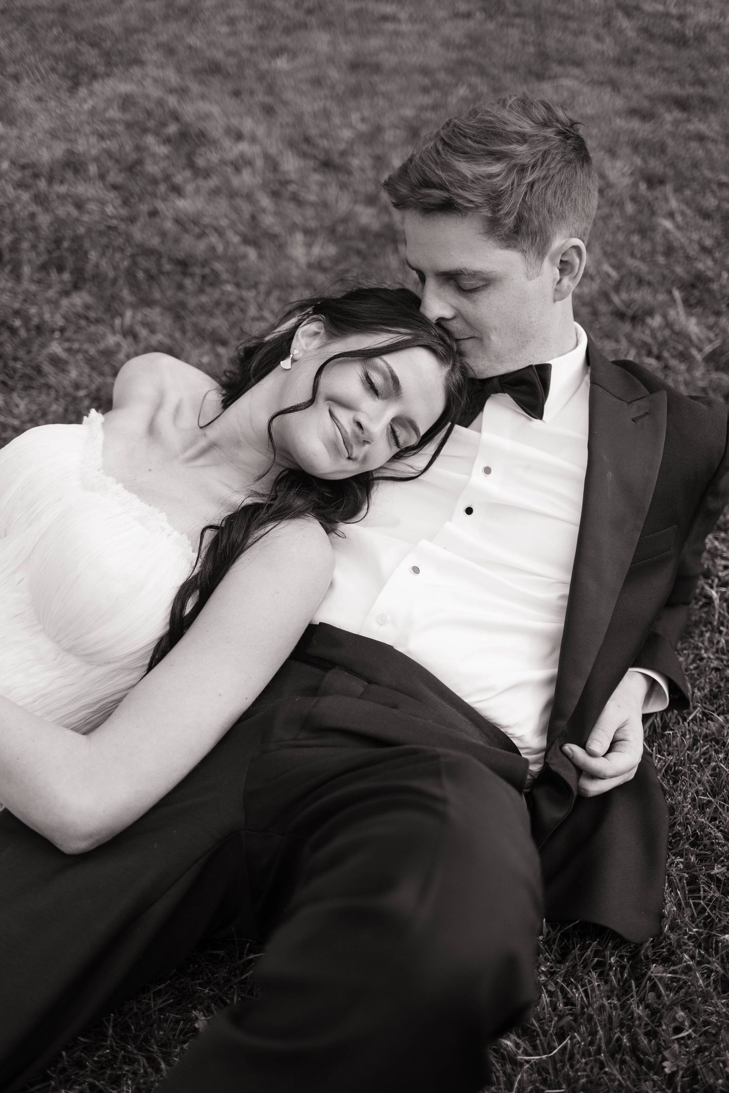 Black and white photo of a bride and groom relaxing on grass, bride resting her head on groom's shoulder.