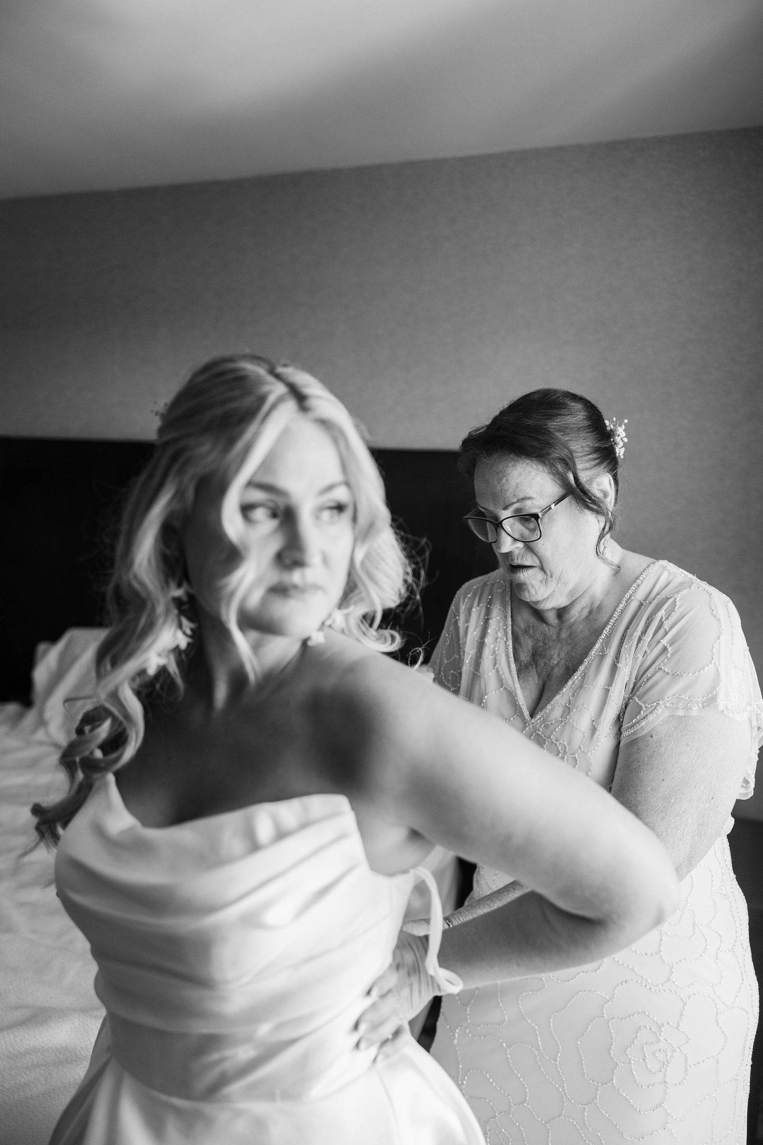 Black and white photo of a bride in a strapless gown being helped into her dress by an older woman in a hotel room.