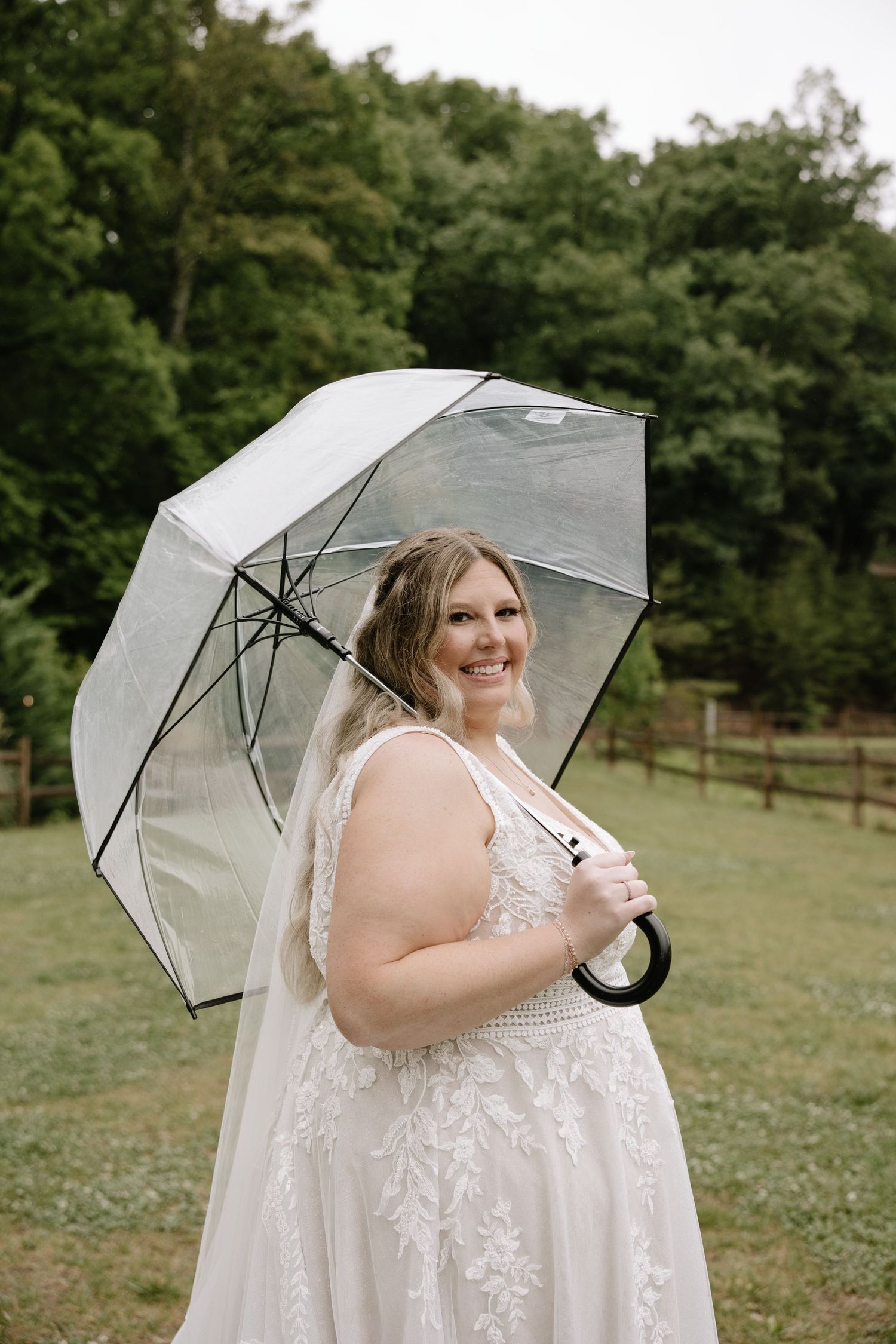 Smiling bride in lace wedding dress holding a clear umbrella outdoors on a green lawn with trees in the background.