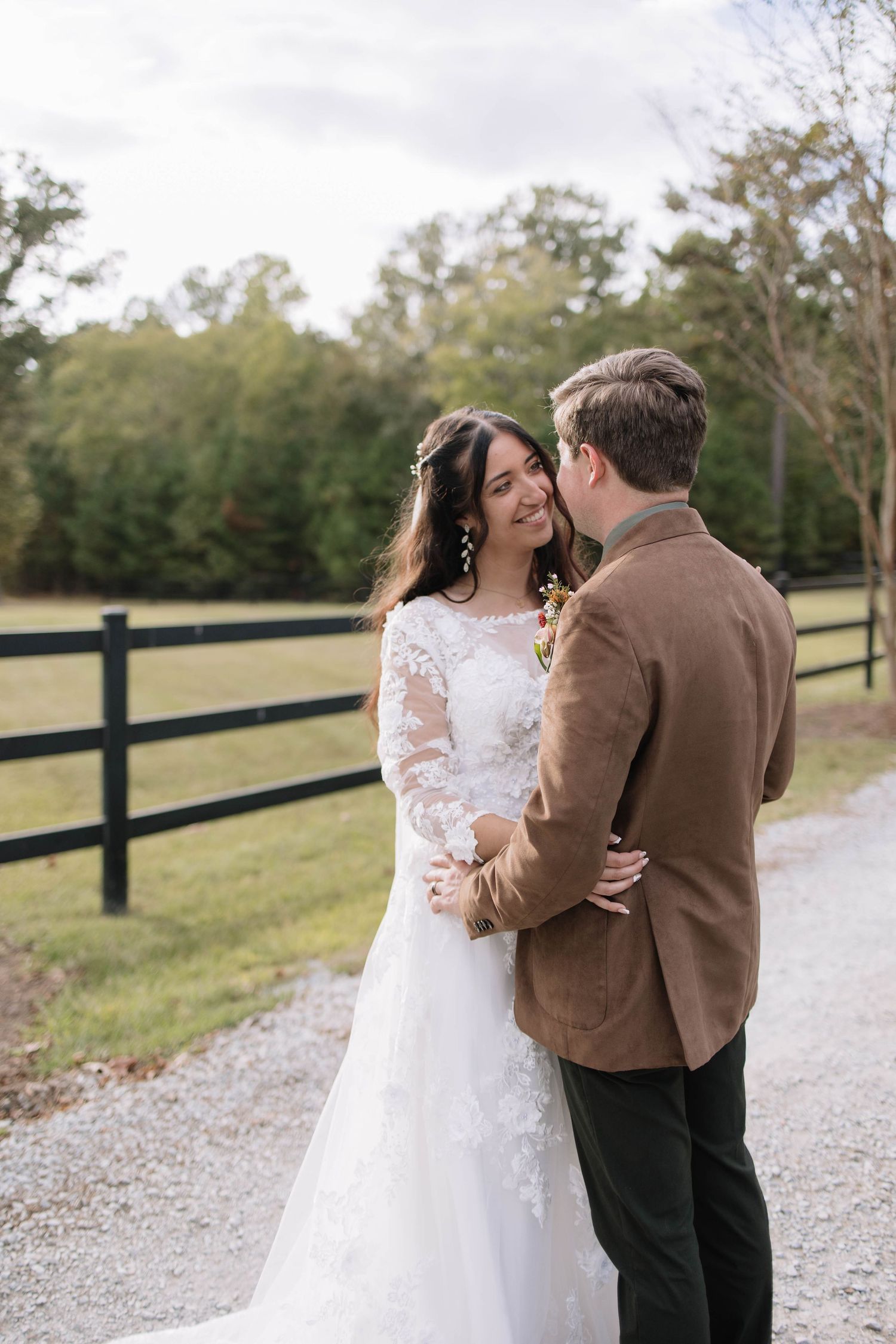 Couple in wedding attire walks along a gravel path bordered by black fencing.