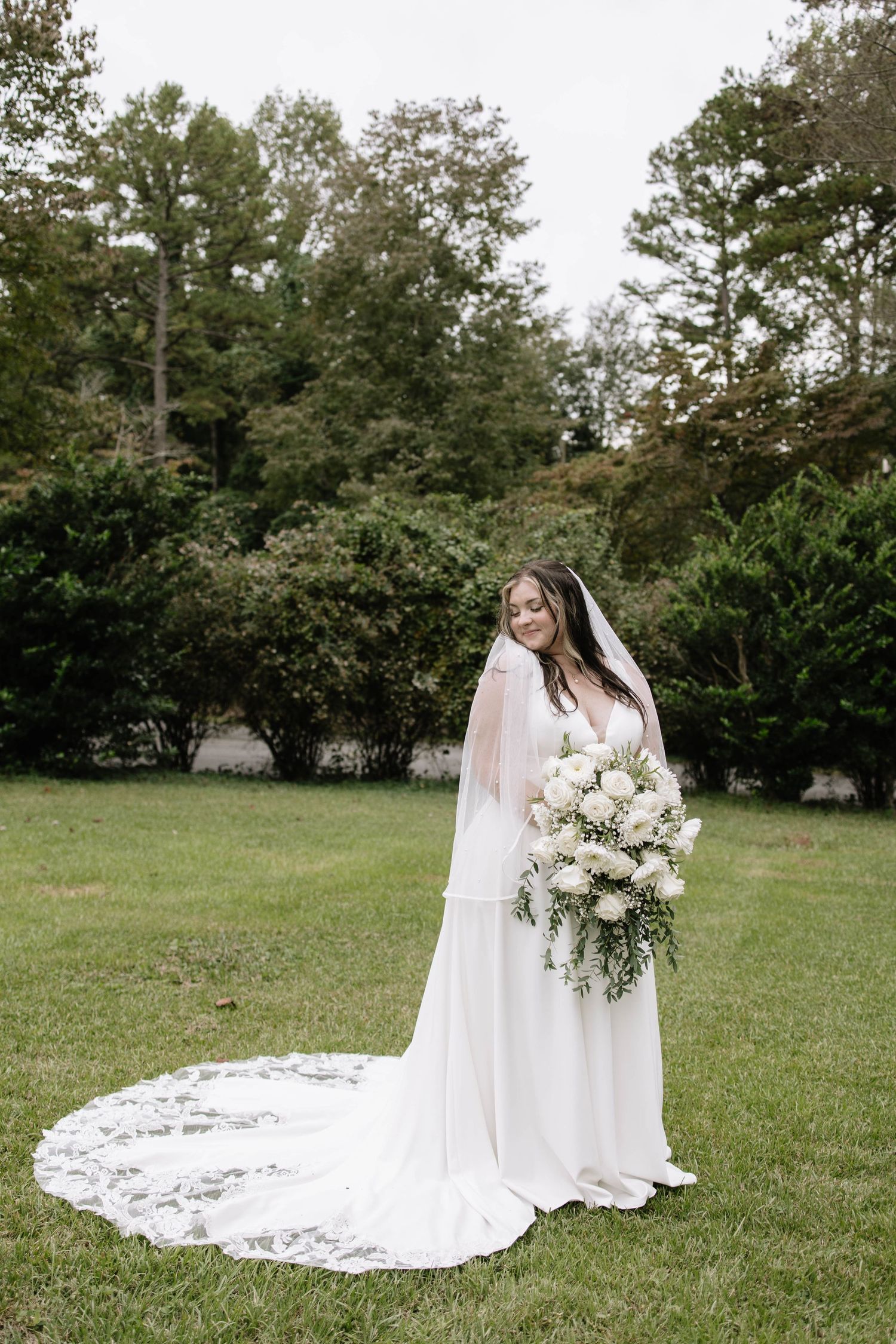 Bride in elegant white gown holds bouquet while standing in outdoor garden setting.