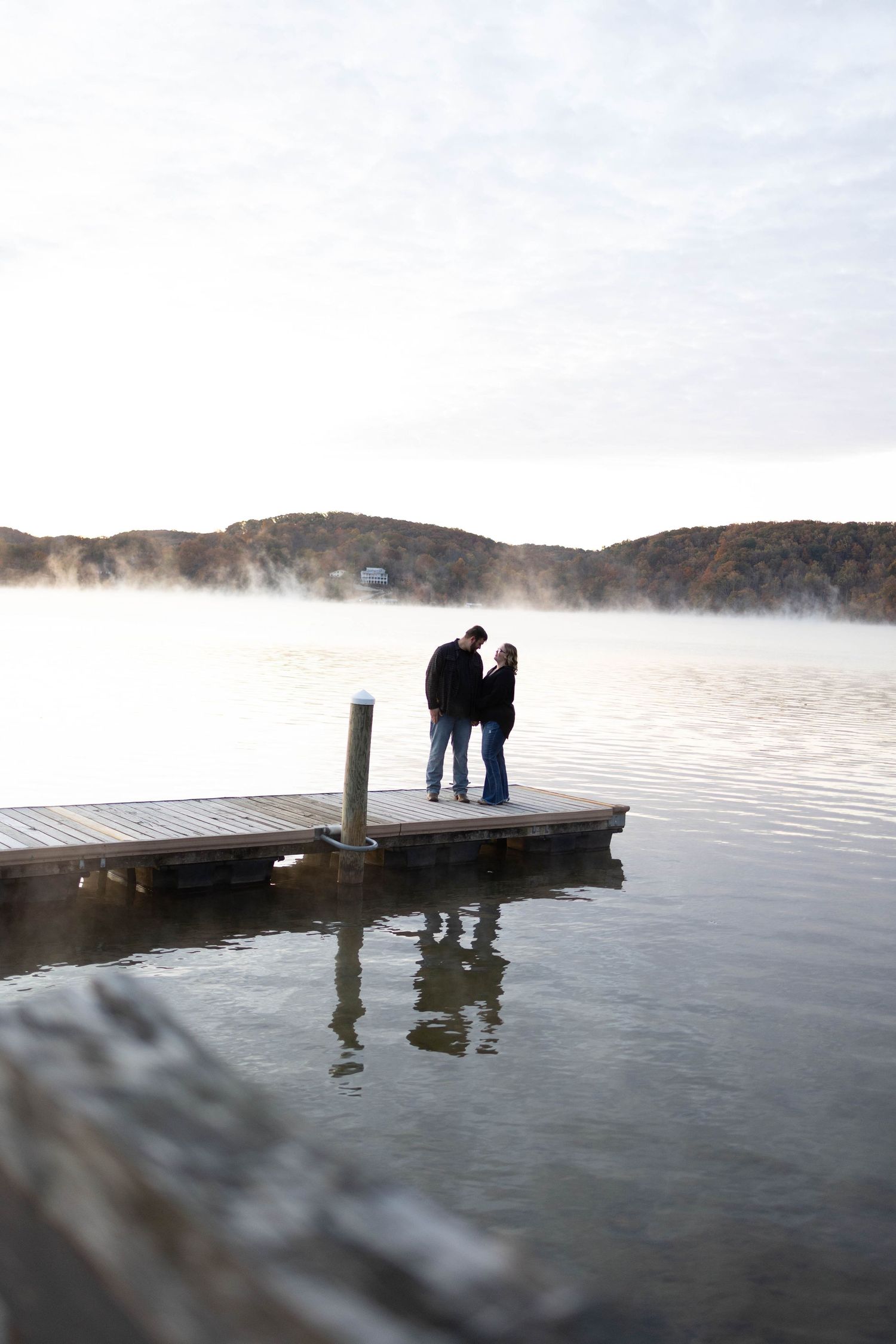 Two silhouettes standing on a wooden dock over a foggy lake with mountains in background.