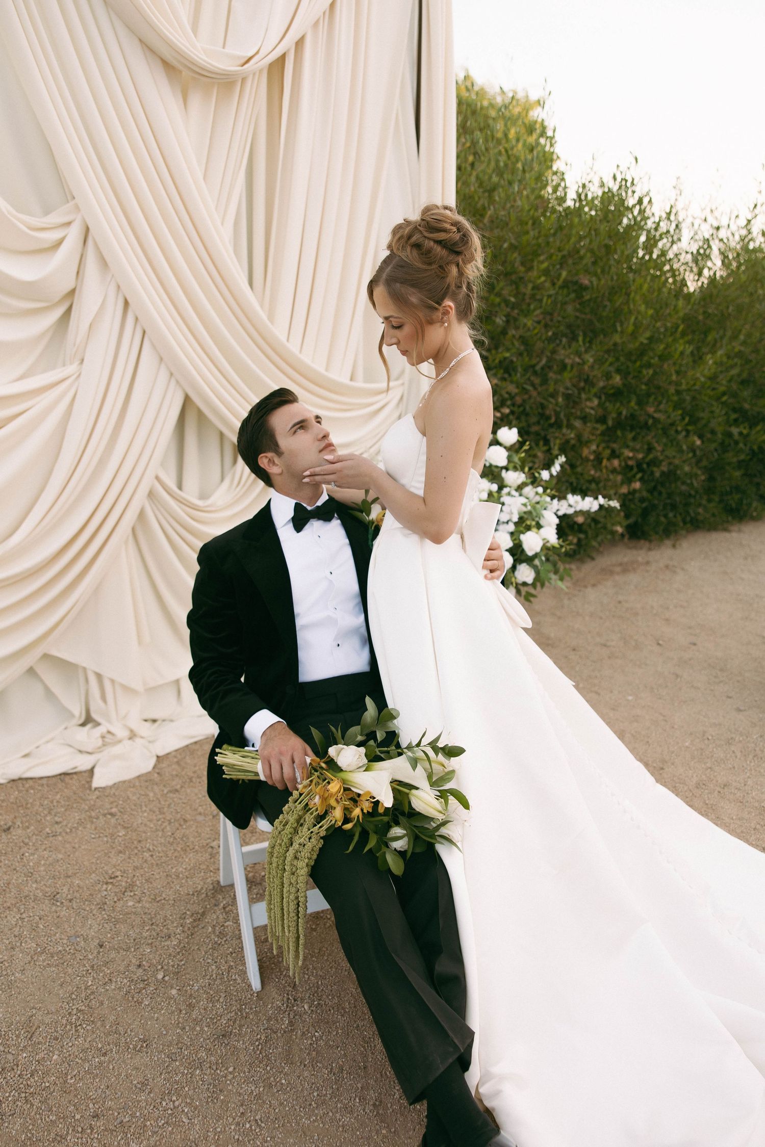 Bride in white gown holds bouquet beside groom in black tuxedo seated before elegant cream draping outdoors.