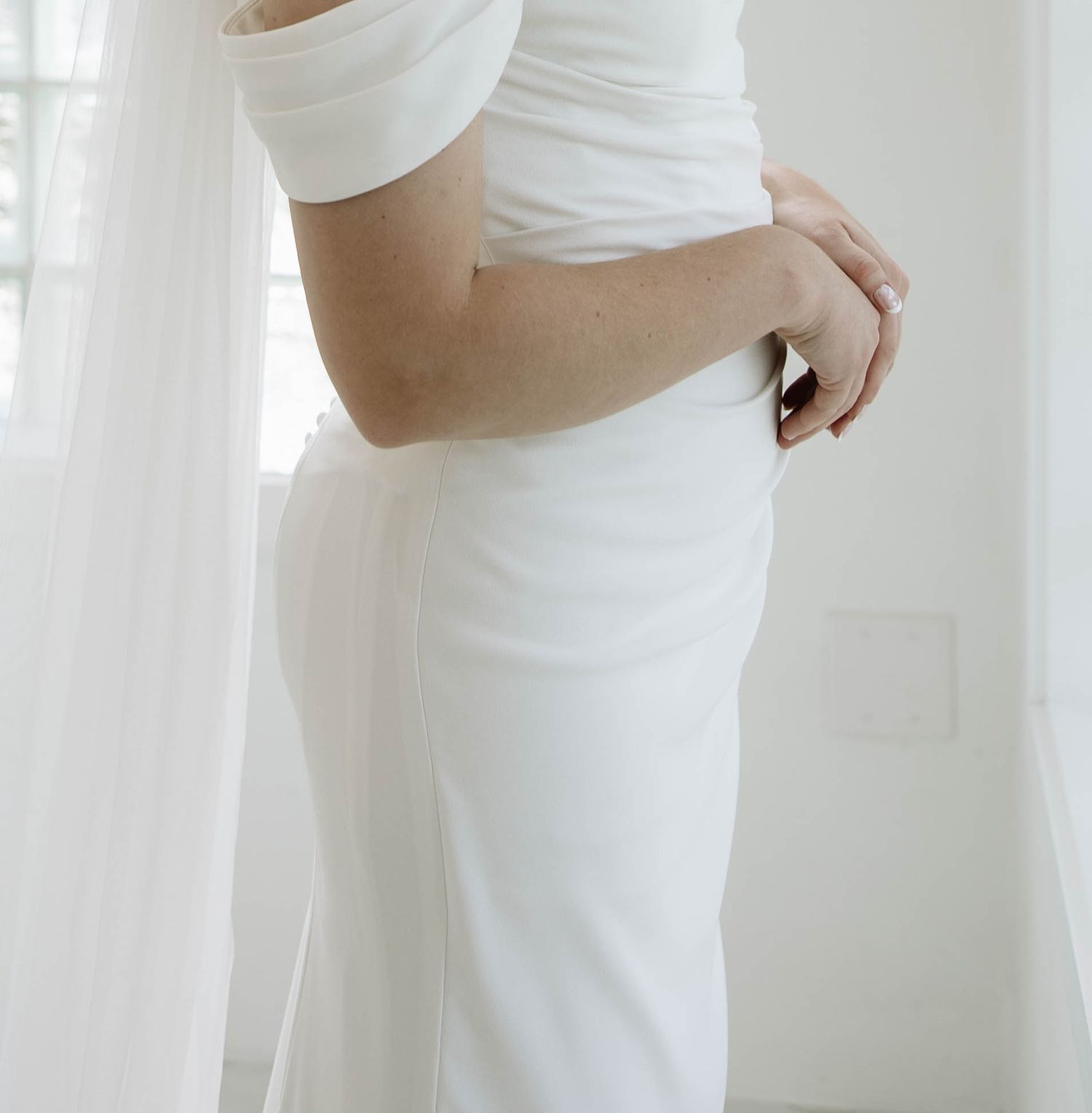 A person wearing an elegant white dress poses gracefully against a bright, airy background with sheer curtains.