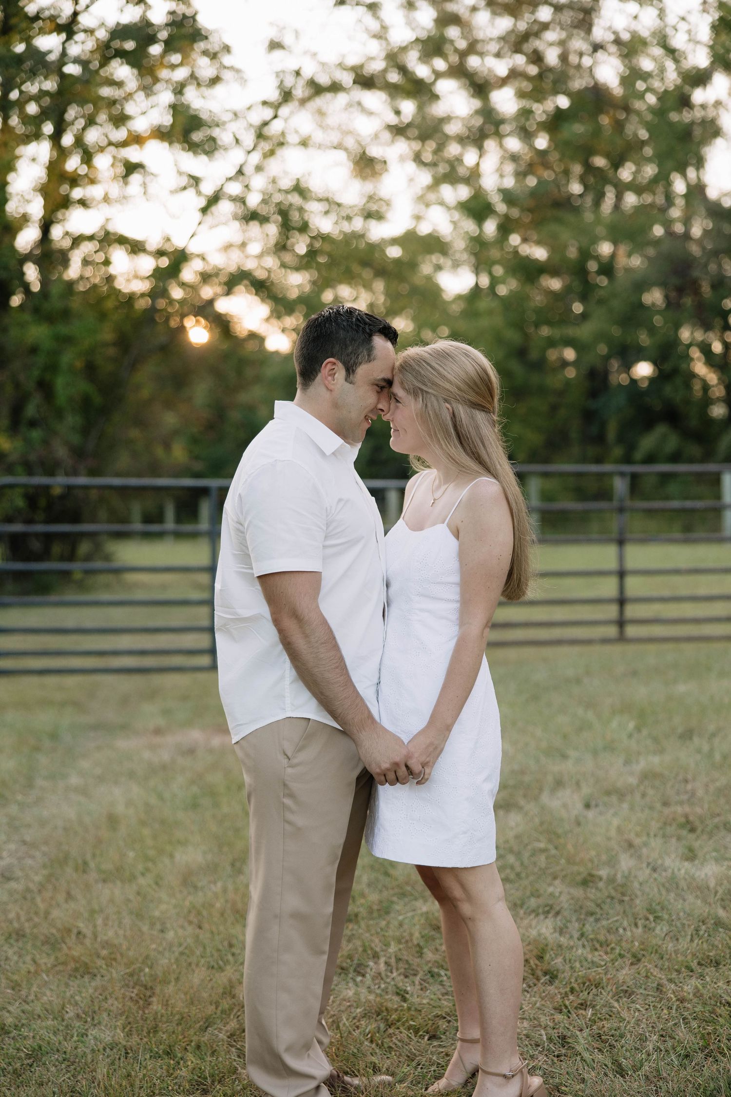 A couple in white outfits sharing an intimate moment in a grassy field at sunset.