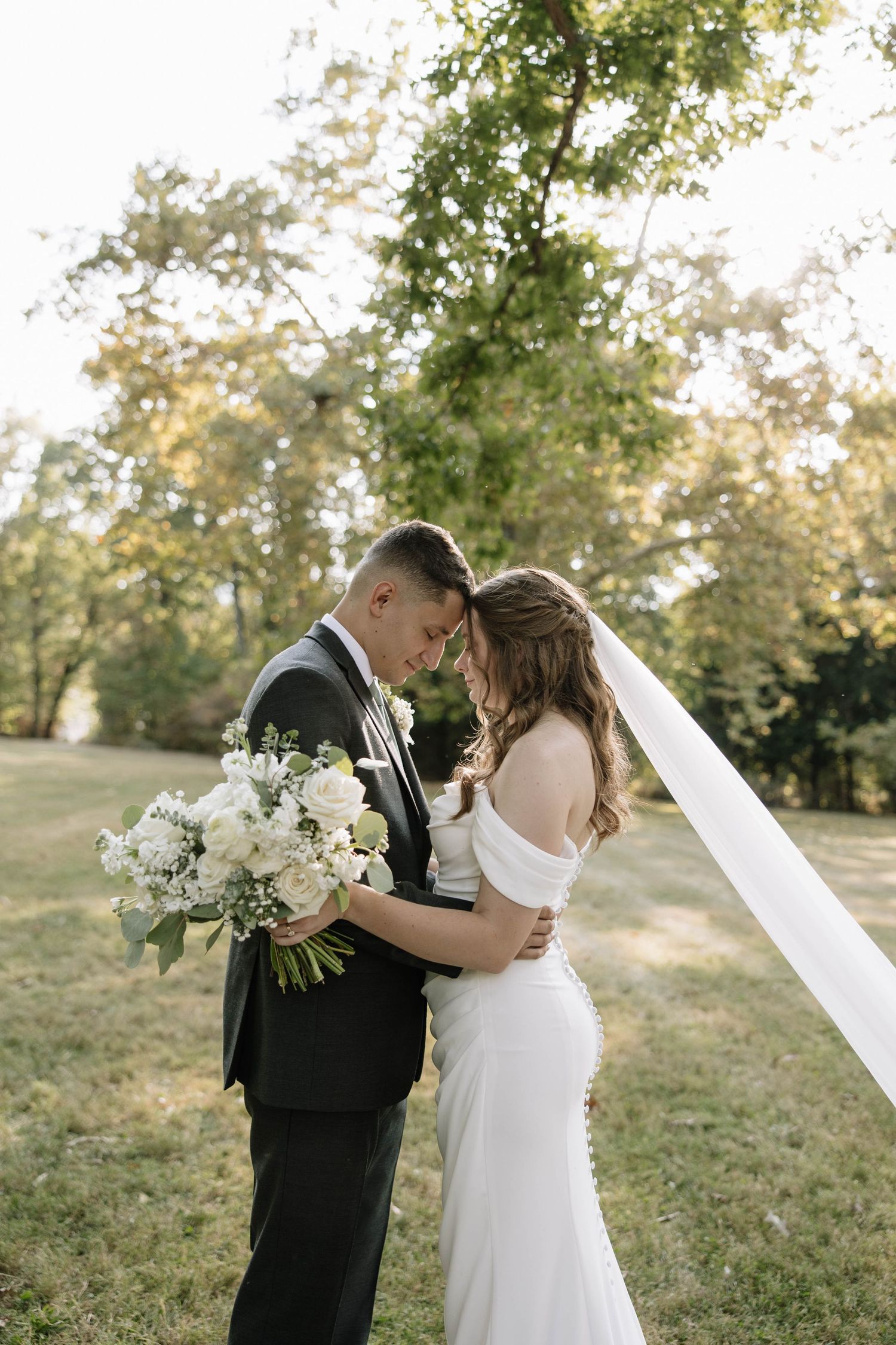 A romantic wedding couple embraces on a sunlit grassy hillside with white floral bouquet and flowing veil.