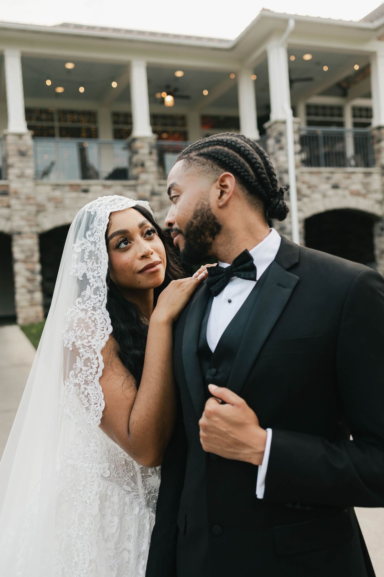 Elegant bride in lace veil and groom in black tuxedo sharing a romantic moment outside a stone venue.
