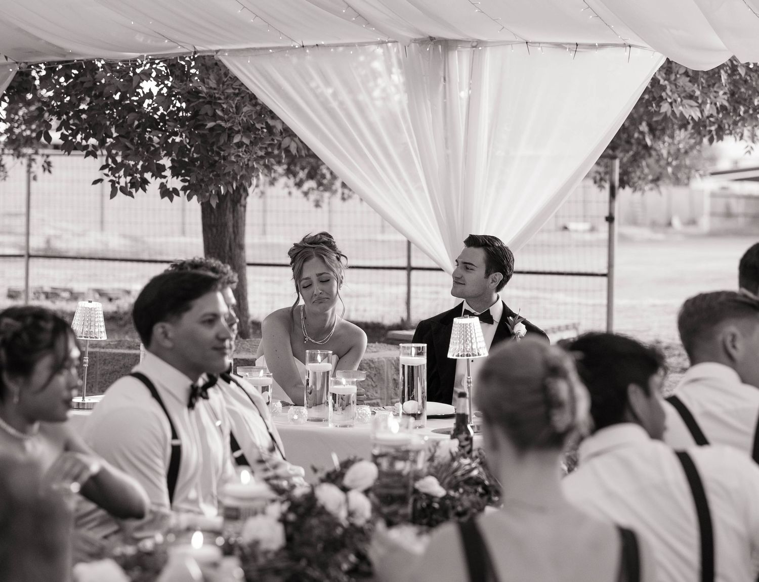 Black and white wedding reception photo of smiling bride and groom seated at head table under elegant draped tent.