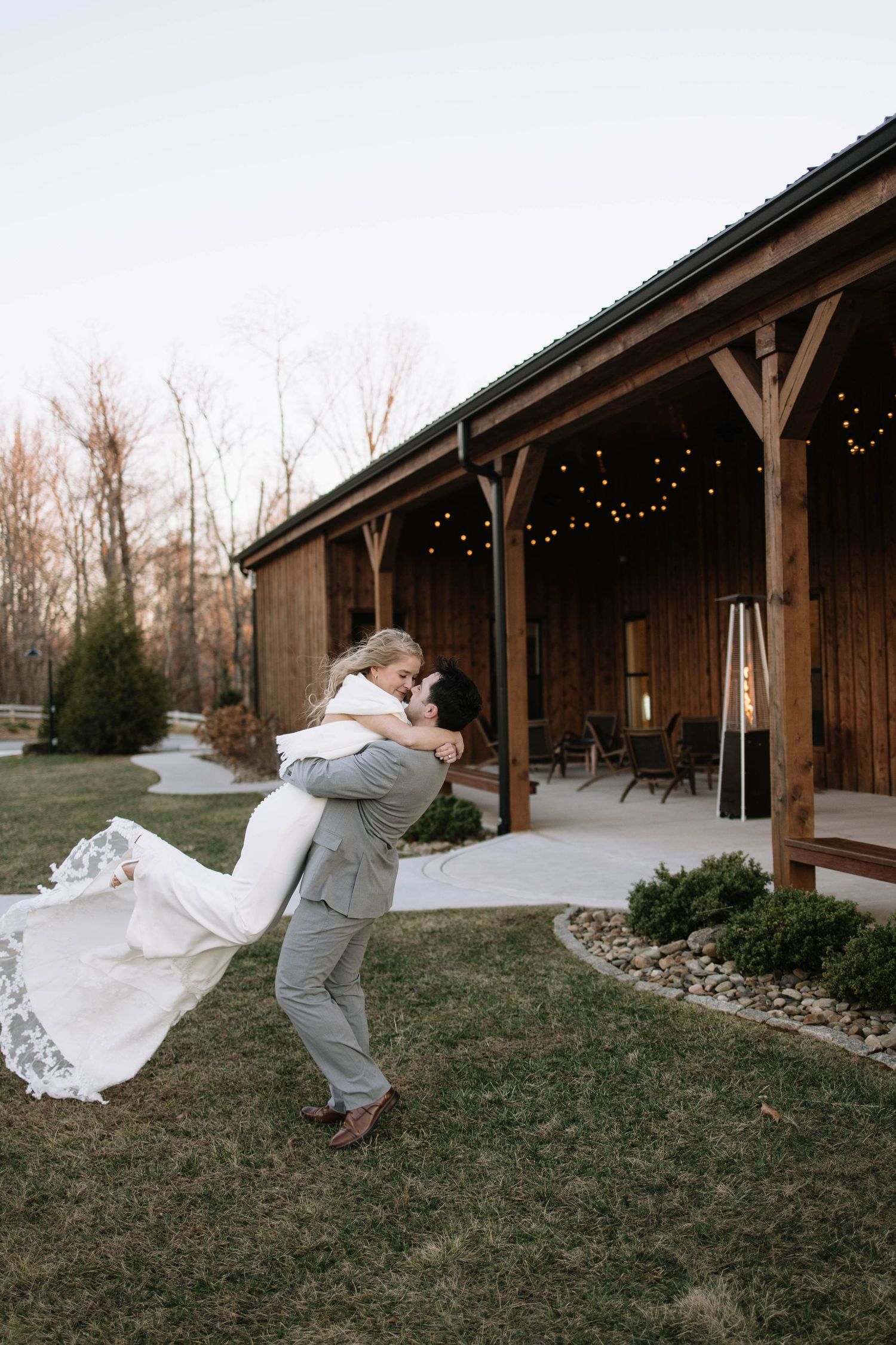 Wedding couple playfully poses with flowing veil against rustic wooden barn backdrop at sunset.