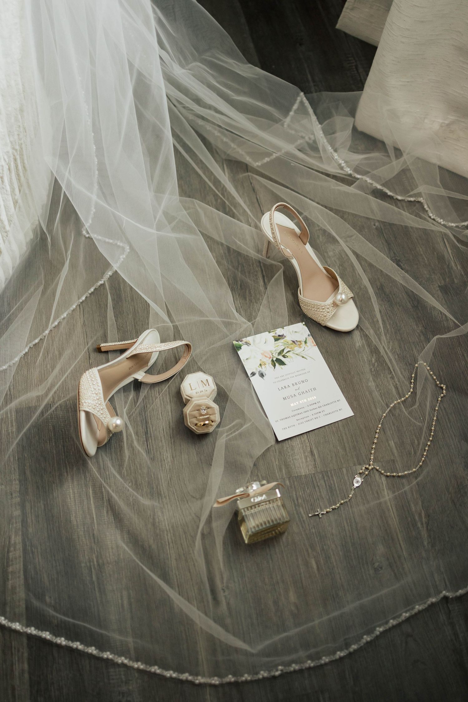 Bridal flat lay with ivory heels, veil, wedding invitation, perfume bottle, and delicate necklace on wood floor.